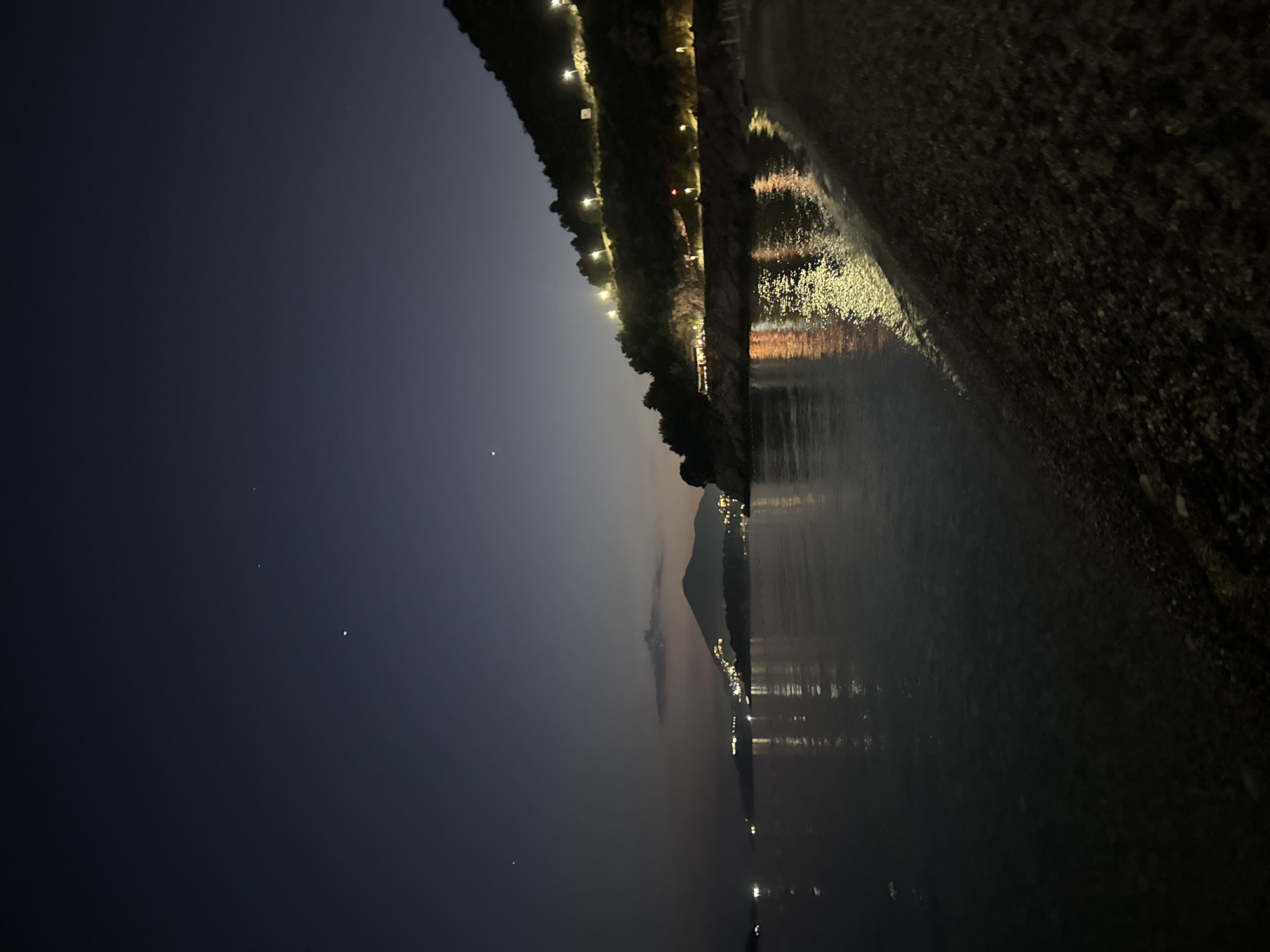The image shows a night view of Kamenovo, Montenegro. A beach extends into the water on the right. Lights line the shore and reflect in the water. A dark mountain is visible in the distance. The sky is a gradient of dark blues and grays, with a few visible stars.