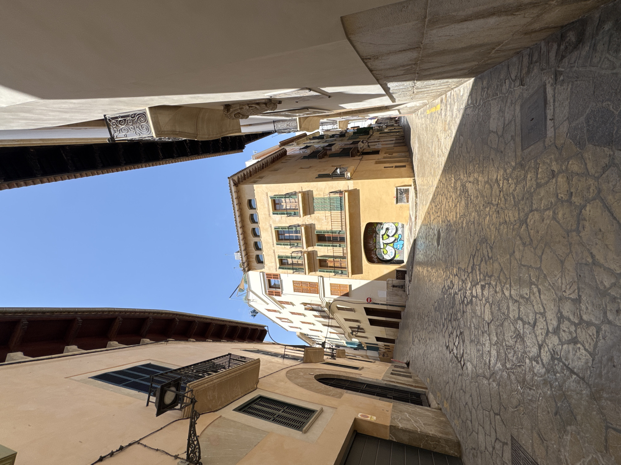 The image shows a narrow, cobblestone street in Palma de Mallorca, Spain. The street is lined with tall, old buildings made of stucco and stone, with balconies and wooden shutters. Sunlight streams down, casting shadows on the uneven pavement. Graffiti is visible on the lower portion of a yellow building on the right, depicting colorful cartoon characters. The sky above is a clear, bright blue. No people are visible in the frame.