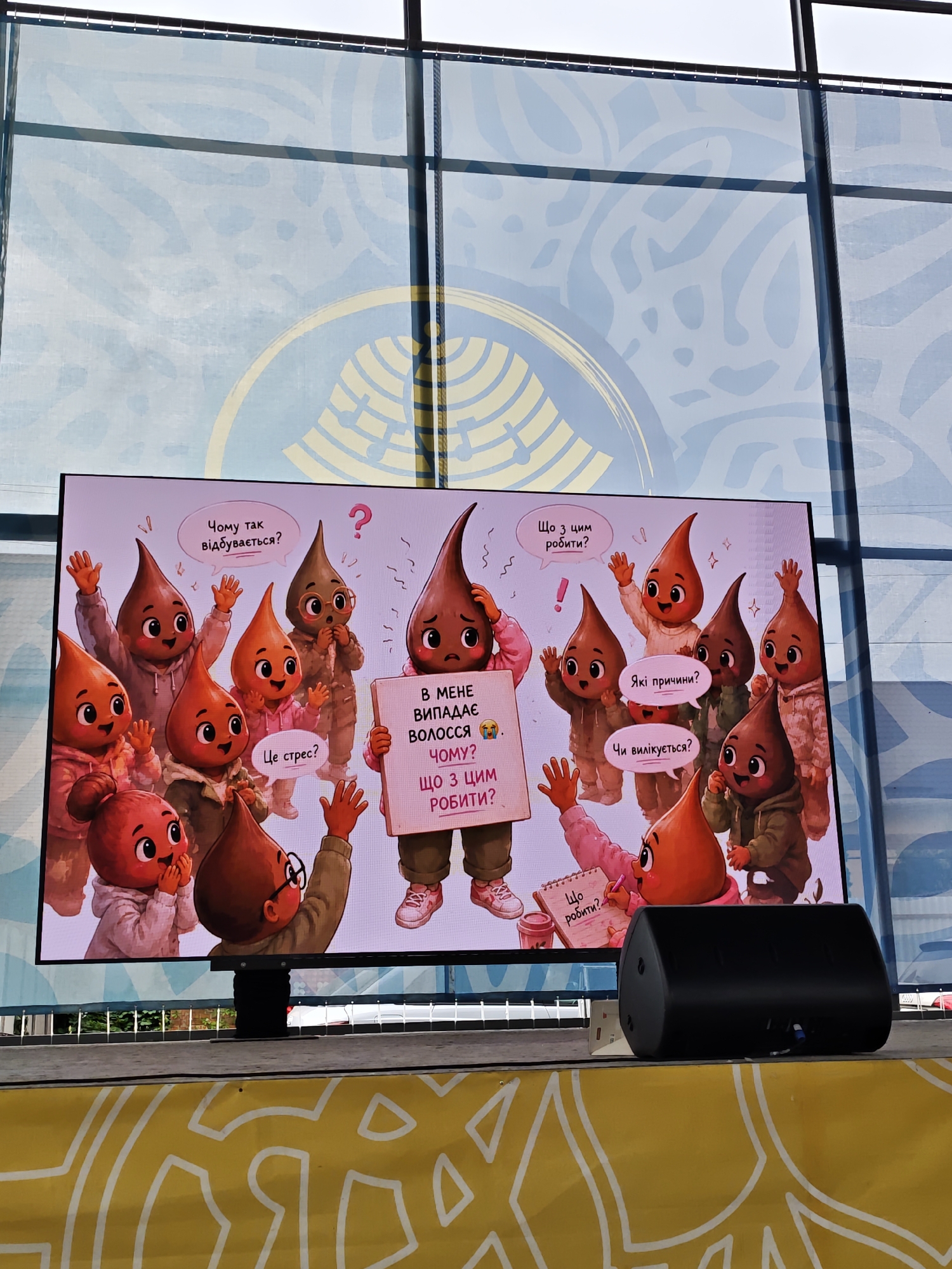 The image depicts an outdoor event setting in Vinnytsia, Ukraine, featuring a large digital screen displaying a colorful cartoon illustration. The illustration centers on numerous anthropomorphic teardrop-shaped characters in shades of brown, orange, and red, each with distinct facial features, some wearing hooded sweatshirts or glasses. A central brown character holds a white sign with Ukrainian text that translates to "MY HAIR IS FALLING OUT WHY? WHAT TO DO ABOUT IT?". Surrounding characters engage in dialogue via speech bubbles, posing questions in Ukrainian such as "Why is this happening?", "Is it stress?", "What to do about it?", "What are the reasons?", and "Will it be cured?".
Below the screen, a black stage monitor speaker is positioned on a dark grey stage or platform. The screen itself is mounted on a visible black stand. In the background, behind the screen, is a large windowed structure covered with a light blue fabric or banner featuring a subtle, lighter blue abstract pattern. A bright, overcast sky is visible above this structure. A thick dark vertical beam and a thinner rope or cord are present on the right side, indicating structural elements. The immediate foreground shows a yellow ground covering or banner adorned with a prominent white abstract or geometric pattern. Green foliage and a partial view of a white vehicle are discernible behind the stage platform in the distance.