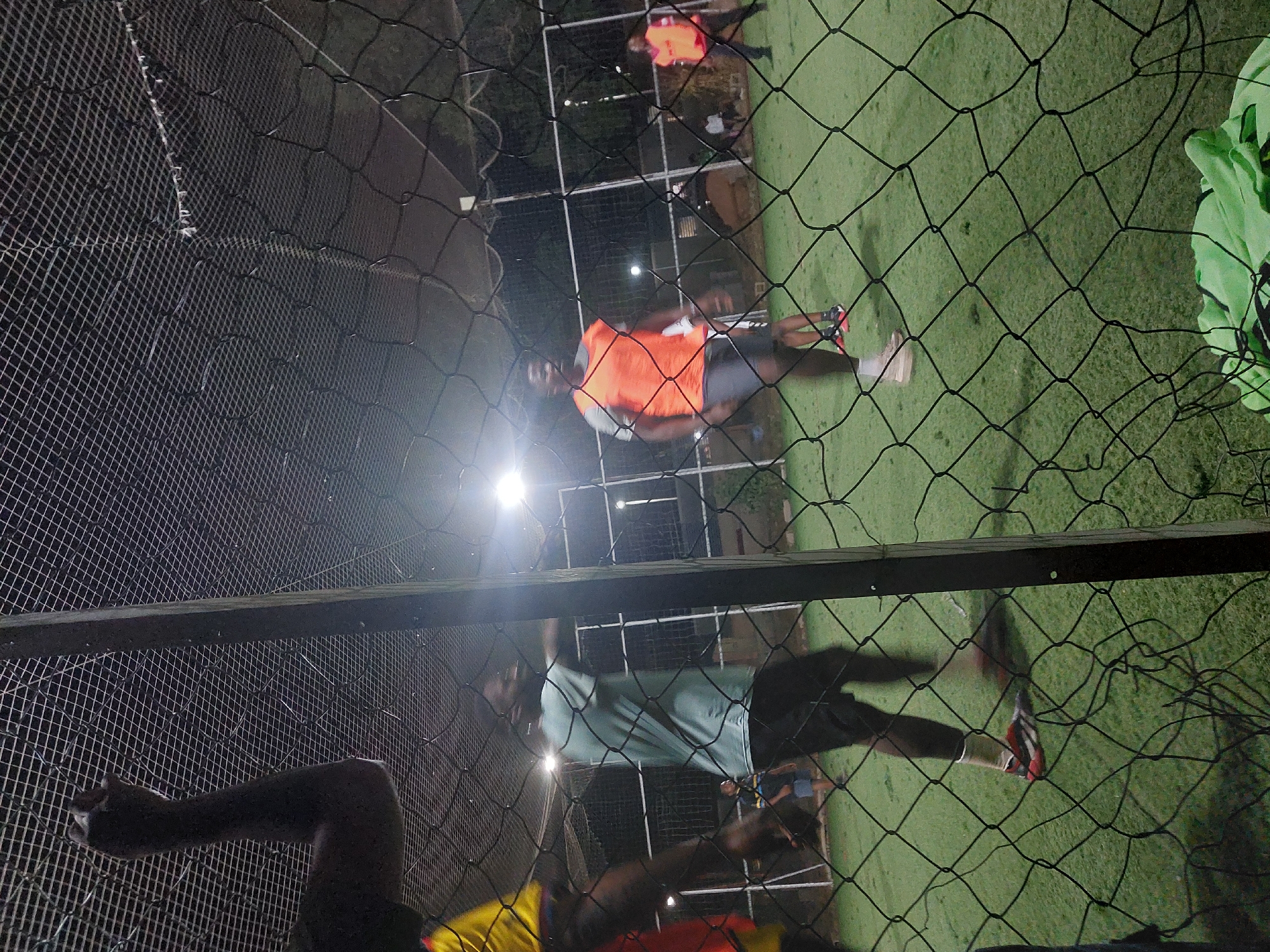 This image depicts an outdoor artificial turf sports field, likely a football or futsal pitch, illuminated at night in Bar Jirgi Summa, Nigeria. The scene is viewed through a prominent black mesh net that covers the foreground, with a finer mesh net visible above.
In the top-left foreground, a dark-skinned arm and shoulder, wearing a yellow garment, is partially visible, seemingly resting on or reaching towards the enclosure netting. To the immediate right, a dark vertical structural post of the enclosure is present.
On the green artificial turf field, multiple dark-skinned male individuals are engaged in activity. In the mid-ground, a male wearing a light blue/green t-shirt, dark shorts, white socks, and red/black athletic shoes stands with his body angled towards the right, appearing in motion. To his right, another male wears an orange bib over a light grey t-shirt, dark shorts, white socks, and light-colored athletic shoes, also appearing in motion. Further into the background, at least one additional person wearing an orange bib is visible, alongside other blurry figures suggesting active play.
The field is brightly lit by several overhead floodlights, causing reflections and glare. White goalposts or field boundary markers are visible in the background. Beyond the field enclosure, dark foliage, possibly trees or shrubs, are discernible. In the bottom-right corner, a crumpled green piece of fabric, possibly clothing or gear, is partially visible on the ground.