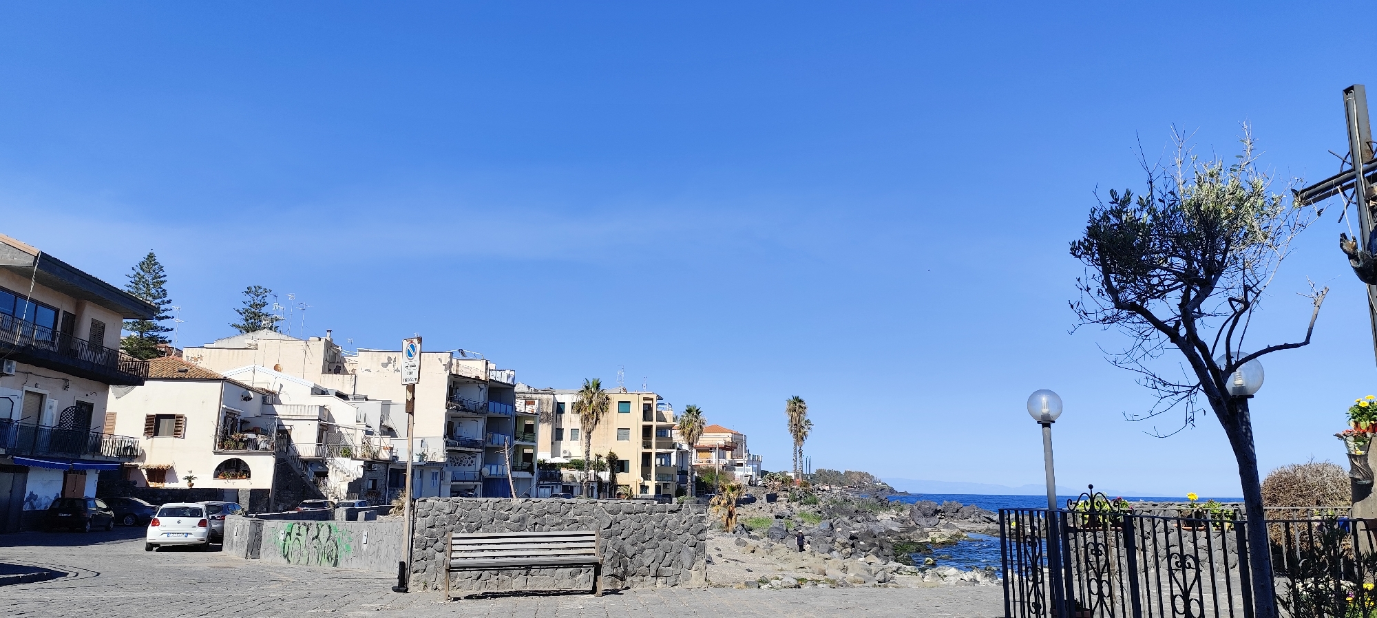 The image depicts a coastal scene in Aci Castello, Italy, under a clear blue sky. Buildings with varying architectural styles line the street on the left. A bench sits atop a stone wall on the right, along with palm trees and other vegetation. The foreground features a stone-paved area, and a black wrought iron fence. To the right, a lamp post and a tree stand near the fence. Beyond the rocky coastline, the sea stretches to the horizon, where a distant landmass is visible. The scene seems calm.