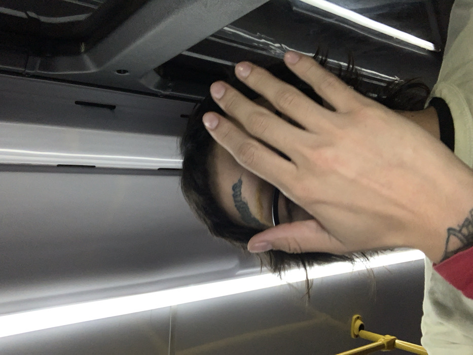 The image depicts an individual partially obscuring their face with their right hand while inside a public transport vehicle in Buenos Aires, Argentina. The environment is characterized by an interior featuring overhead fluorescent strip lighting and grey structural paneling, with a yellow vertical grab pole visible on the left. The subject, of indeterminate gender, has dark, medium-length hair. A dark, stylized tattoo or marking reading "agresor" is visible on their left eyebrow, which also shows a yellowish tint. Part of a thin glasses frame is visible below the hand. The individual wears a light-colored t-shirt with a dark collar. A red sleeve cuff is visible on the left wrist, below which a dark, intricate tattoo is partially visible. The person's hand is raised, palm facing the camera, covering the majority of the face.