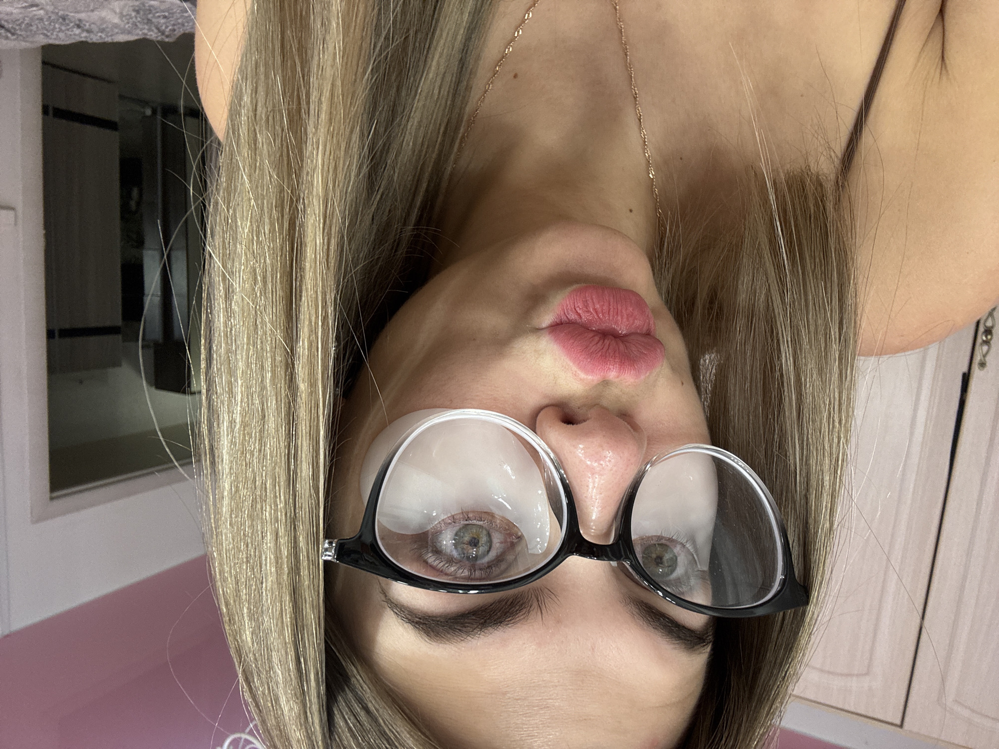 The image depicts a close-up forensic view of an adult female, likely indoors within a private residence in the city of Chaikovsky, Russia. The subject has long, light brown hair styled down, green eyes, and is looking directly at the camera. She wears black-rimmed glasses, underneath which white, crescent-shaped under-eye patches are visible. Her lips are pursed in a "kissing" gesture, accentuated by pink lipstick. A thin, gold-colored chain necklace with small links is visible around her neck, and she appears to be wearing a dark, thin-strapped top.
The background elements include a light-colored wooden wardrobe or cabinet with two doors on the left. The upper right portion shows a pink-colored wall or ceiling section. To the right, a white door frame opens into a darker adjacent room or hallway, revealing a dark door with horizontal light-colored stripes. The bottom right edge of the frame shows a patterned grey and white fabric, consistent with a bedspread or blanket. Artificial lighting illuminates the scene, causing reflections on the glasses. The subject's pose indicates a deliberate action for the photograph.