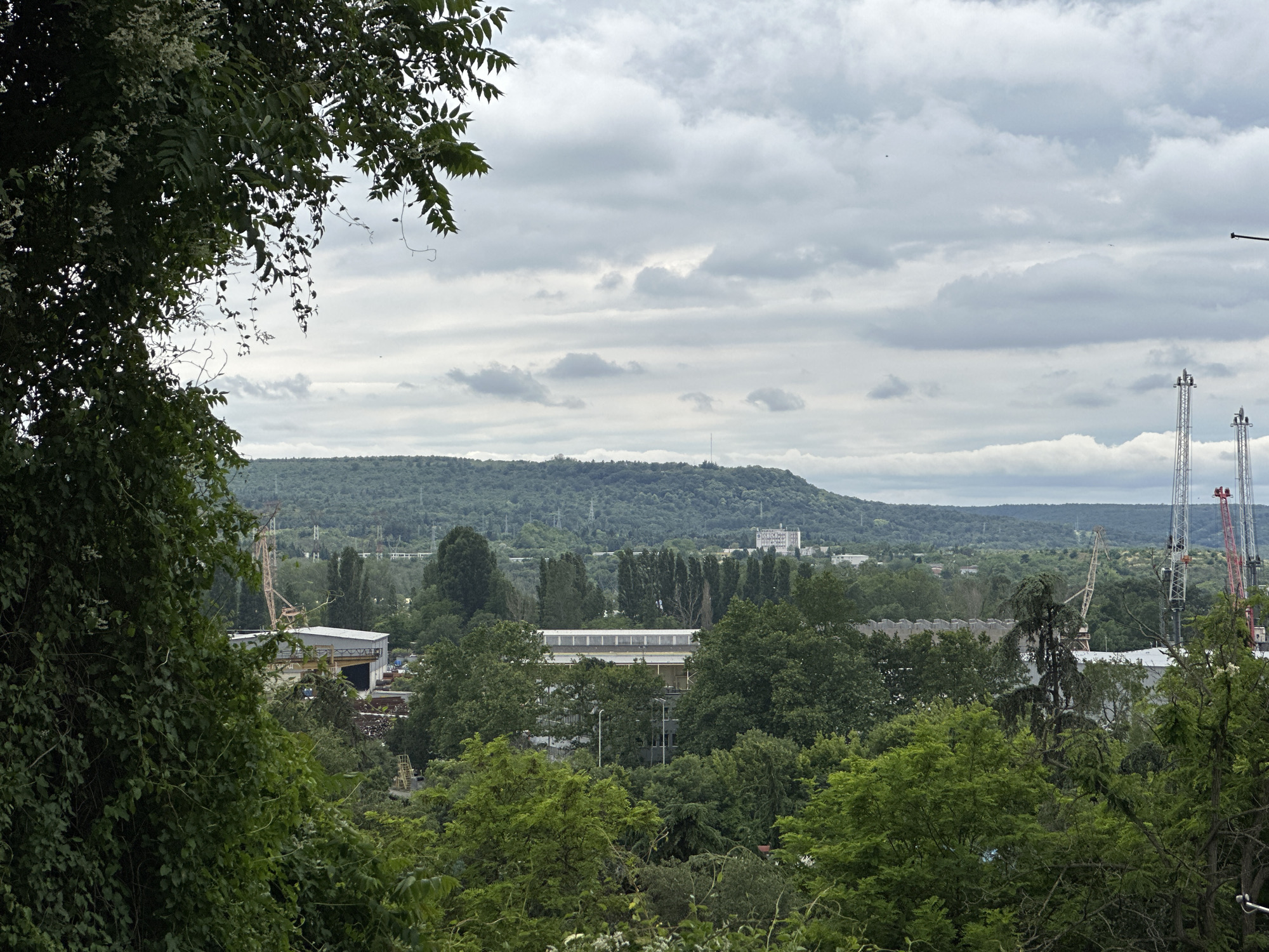A medium shot captures a panoramic view of Varna, Bulgaria, with lush green foliage in the foreground framing a cityscape. The sky is overcast with a mosaic of gray and white clouds. In the middle ground, industrial buildings and infrastructure are visible, including a large gray building with a white roof and numerous windows, surrounded by trees. Several cranes, some red and white, are present, indicating ongoing construction or industrial activity. In the background, a rolling green hill dominates the landscape, dotted with trees and topped with a cluster of buildings. Power lines are also visible across the hills. The scene appears to be a typical urban industrial area with natural elements.