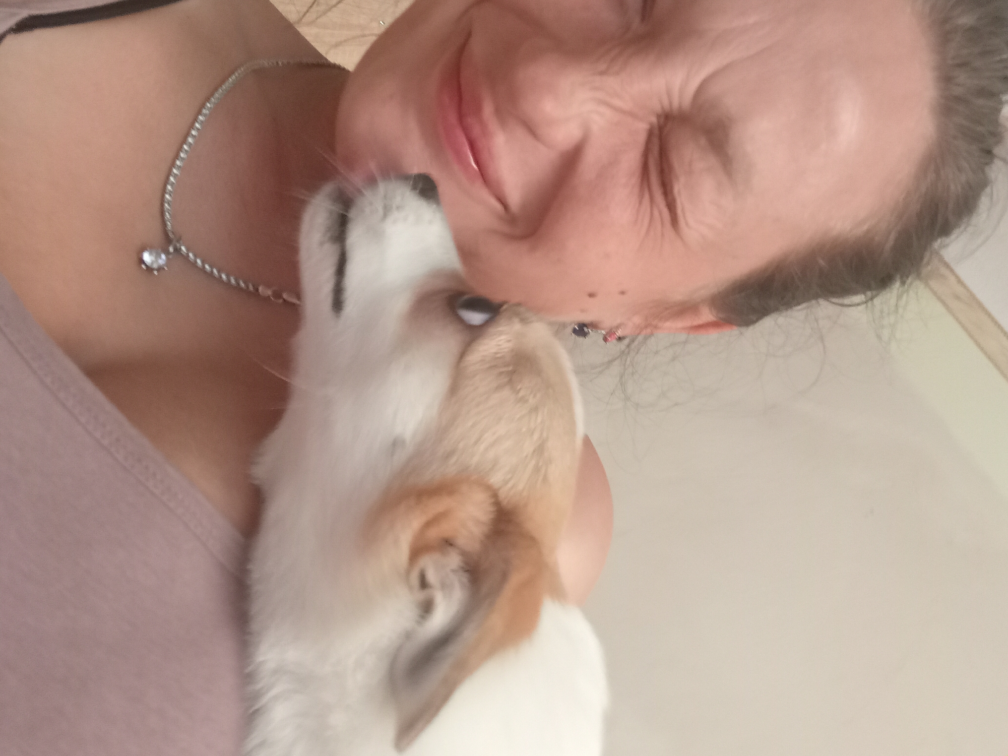 A woman is taking a selfie with a small dog, resting the dog on her shoulder. The woman is smiling with her eyes closed, her face wrinkled in a playful expression. She is wearing a necklace with a sparkling pendant. The dog, a light-colored breed with white and tan fur, is looking towards the camera with its mouth slightly open. The background is a plain, light-colored wall. The image appears to have been taken indoors. The location in the city of Chuhuiv, Ukraine is not discernible from the provided image.