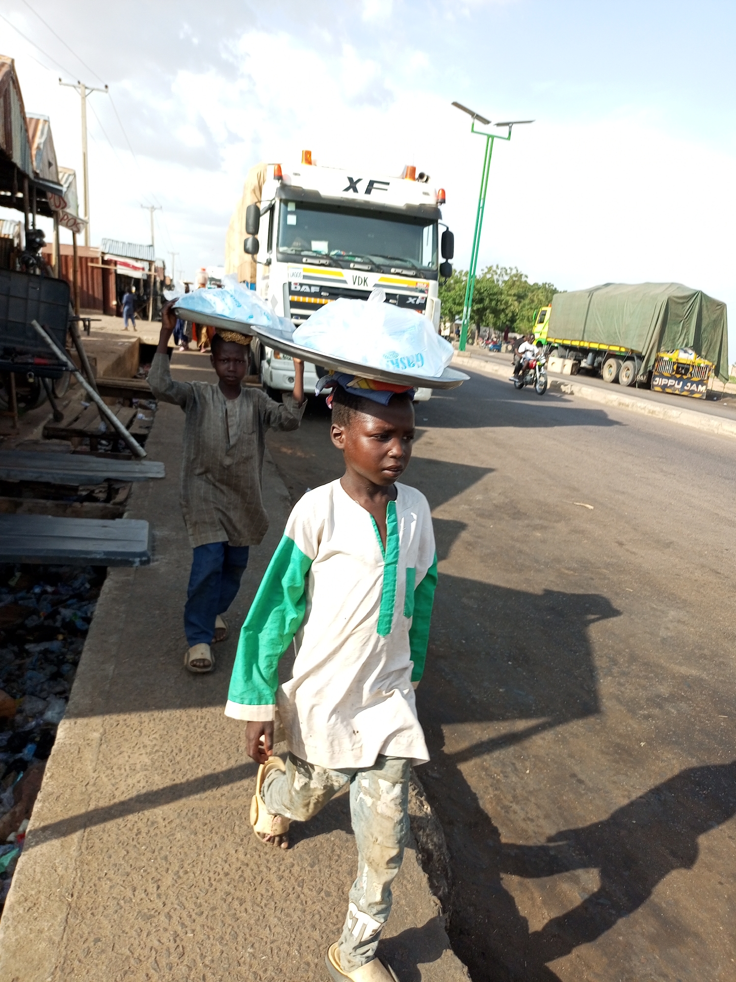 In Gadao, Nigeria, two young boys are seen carrying trays of goods on their heads as they walk along a dusty roadside. The boy in the foreground wears a white and green tunic and patterned trousers, with his right foot in a sandal. Behind him, another boy in a striped tunic and blue trousers also carries a tray. In the background, a white truck with "XF" branding is parked, and a green-covered truck with "JIPPU JAM" written on its side is on the road. The scene depicts a busy commercial area with market stalls and a main road, suggesting a common sight of child vendors in the city.