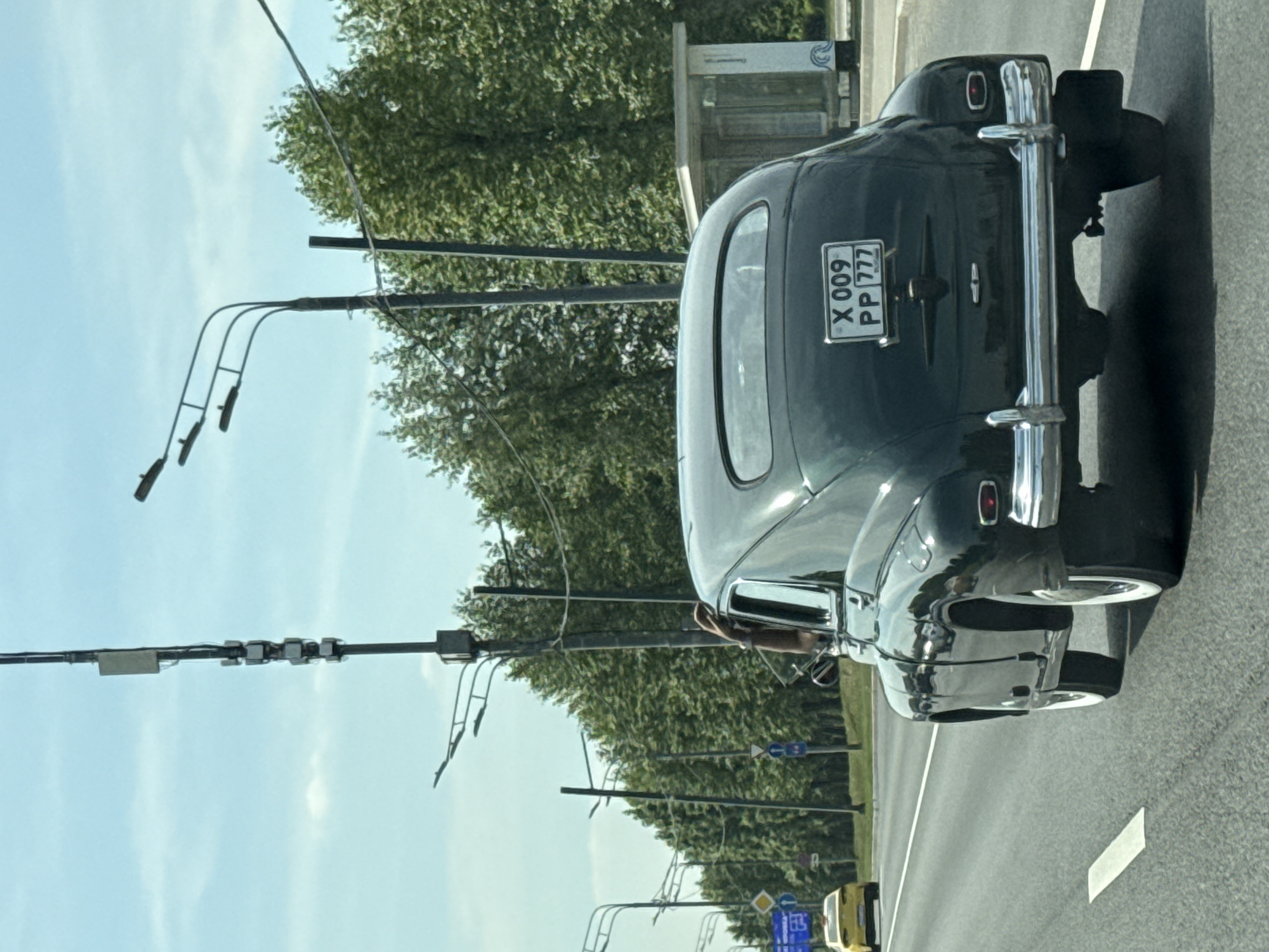 The image depicts a dark green vintage GAZ-M20 "Pobeda" sedan traveling on an asphalt road in Khimki, Russia, on a clear day. The vehicle is observed from the rear, showing its curved roofline, distinctive rear window, and prominent chrome bumper. The Russian license plate "X 009 PP 777" is affixed to the trunk lid, with "RUS" visible below the "777" region code, indicating Moscow and Moscow Oblast. The car features white wall tires. A person's right arm, wearing a watch, is extended out of the open driver's side window.
The road features a solid white line marking the shoulder on the right and a dashed white line separating lanes. In the distance, a yellow taxi is visible in the left lane.
On the left side of the road, multiple poles support streetlights with dual lamp fixtures. A large blue road sign with white text (partially obscured) is visible, along with a round blue sign indicating a mandatory right turn or direction, and another blue sign with a white bicycle symbol.
On the right side, a tall utility pole is equipped with several vertically stacked rectangular modules, possibly antennas or surveillance equipment, and a larger flat rectangular panel higher up. Beyond the vehicle, a small, open-sided bus stop shelter made of white concrete with a decorative wave pattern at its base is present on the roadside. A dense line of green deciduous trees forms a backdrop along the right side of the road. The sky is light blue with sparse white clouds.
