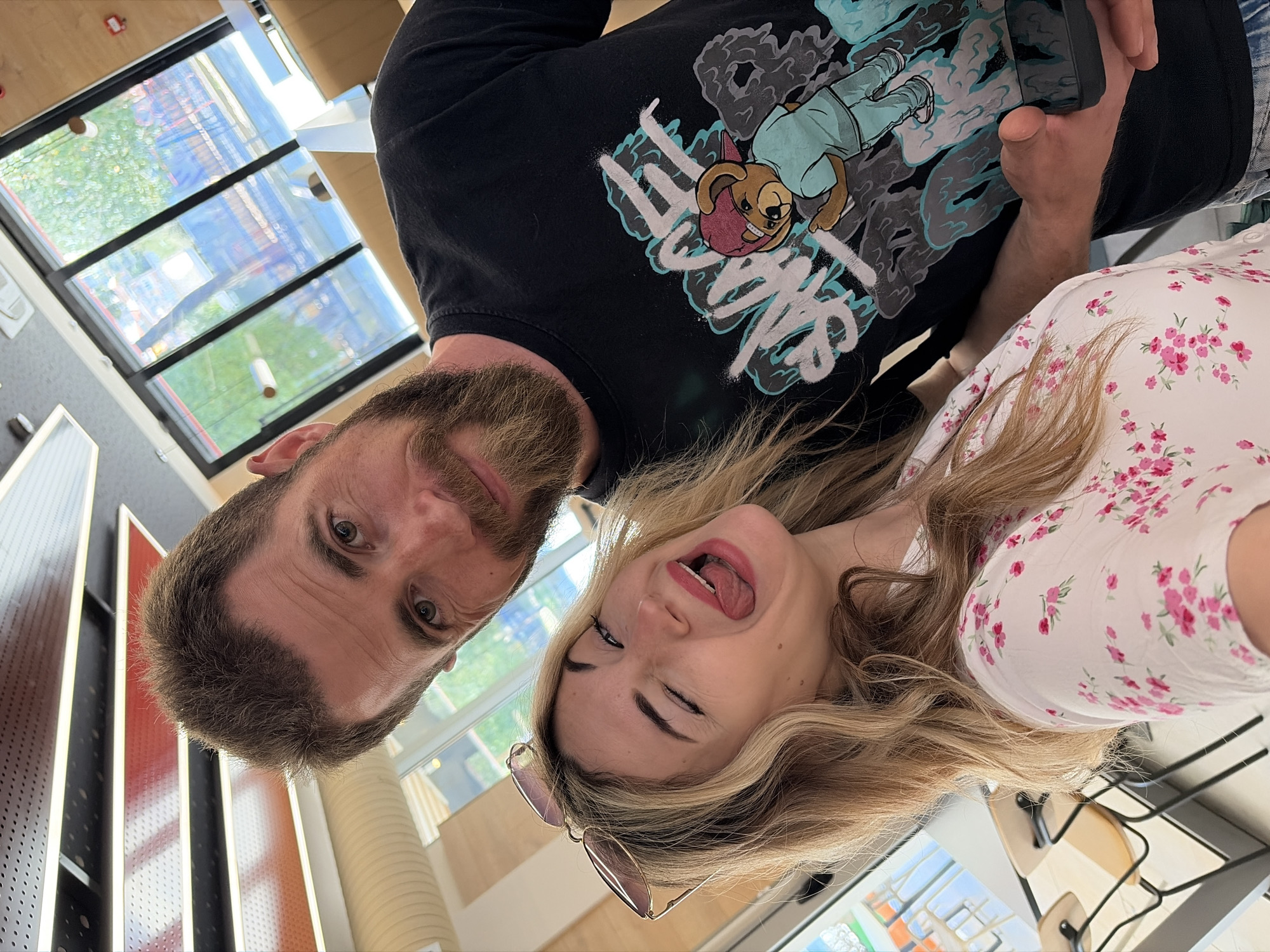 A man and a woman are posing for a selfie. The man has brown hair and a beard. He is wearing a black sweatshirt with a cartoon design. The woman has long blonde hair and is sticking her tongue out and winking. She is wearing a white floral dress and sunglasses. They appear to be inside a building with large windows. Outside the windows, there is a view of buildings and greenery. It is not possible to confirm if this is in Budva, Montenegro, from the image alone.