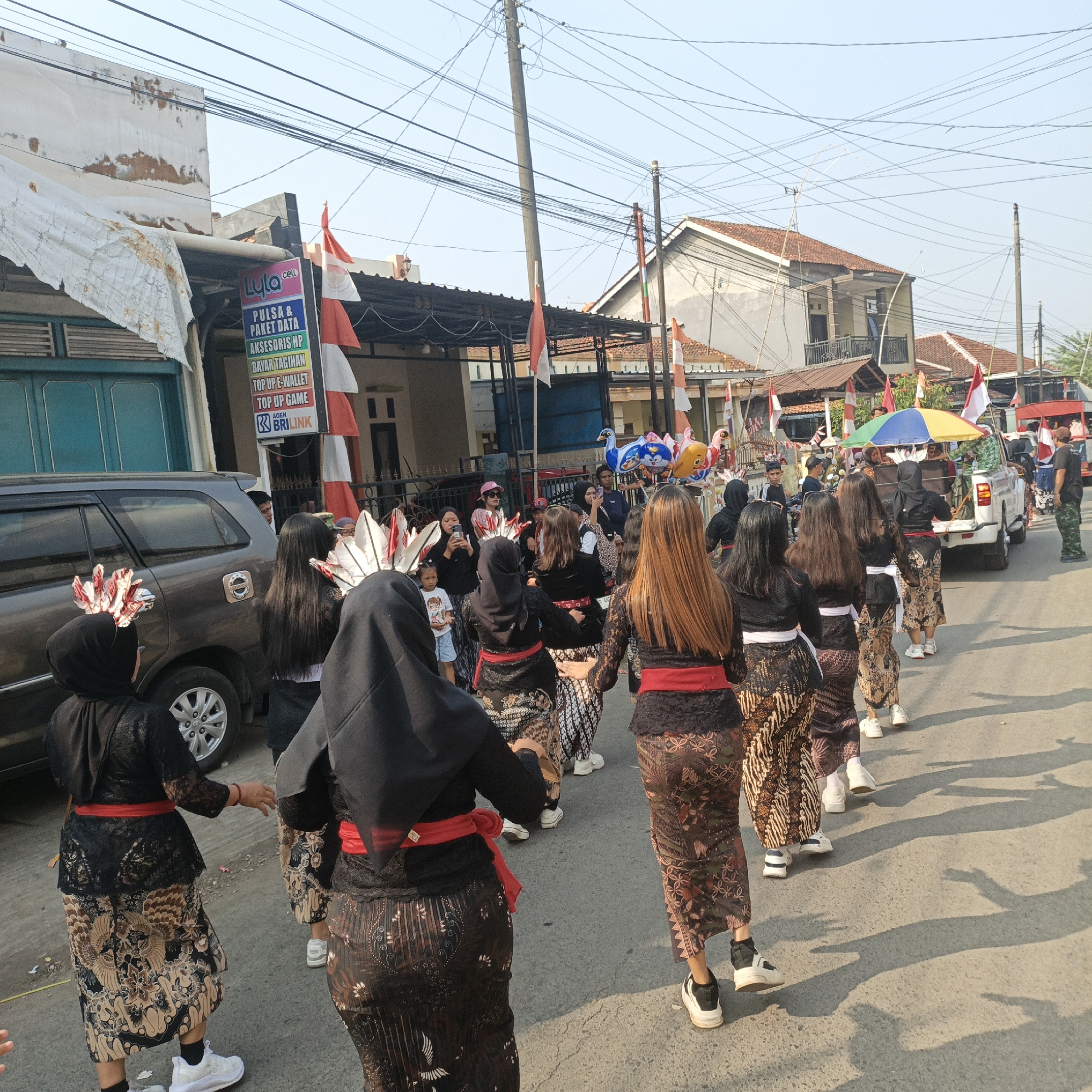 The image depicts a procession on a street in Tanahbaya, Indonesia. A group of individuals, predominantly young women dressed in traditional batik skirts and black lace tops, are walking in a line. Many are wearing red sashes and elaborate headpieces resembling feathers. Some are holding phones, possibly recording the event. In the background, a grey SUV is parked, and a white truck with a colorful umbrella on top is visible. Indonesian flags are displayed on poles along the street, suggesting a national celebration. Buildings with tiled roofs line the sides of the street, and numerous power lines crisscross the sky. The scene appears to be taking place during the daytime, with sunlight casting shadows on the asphalt.