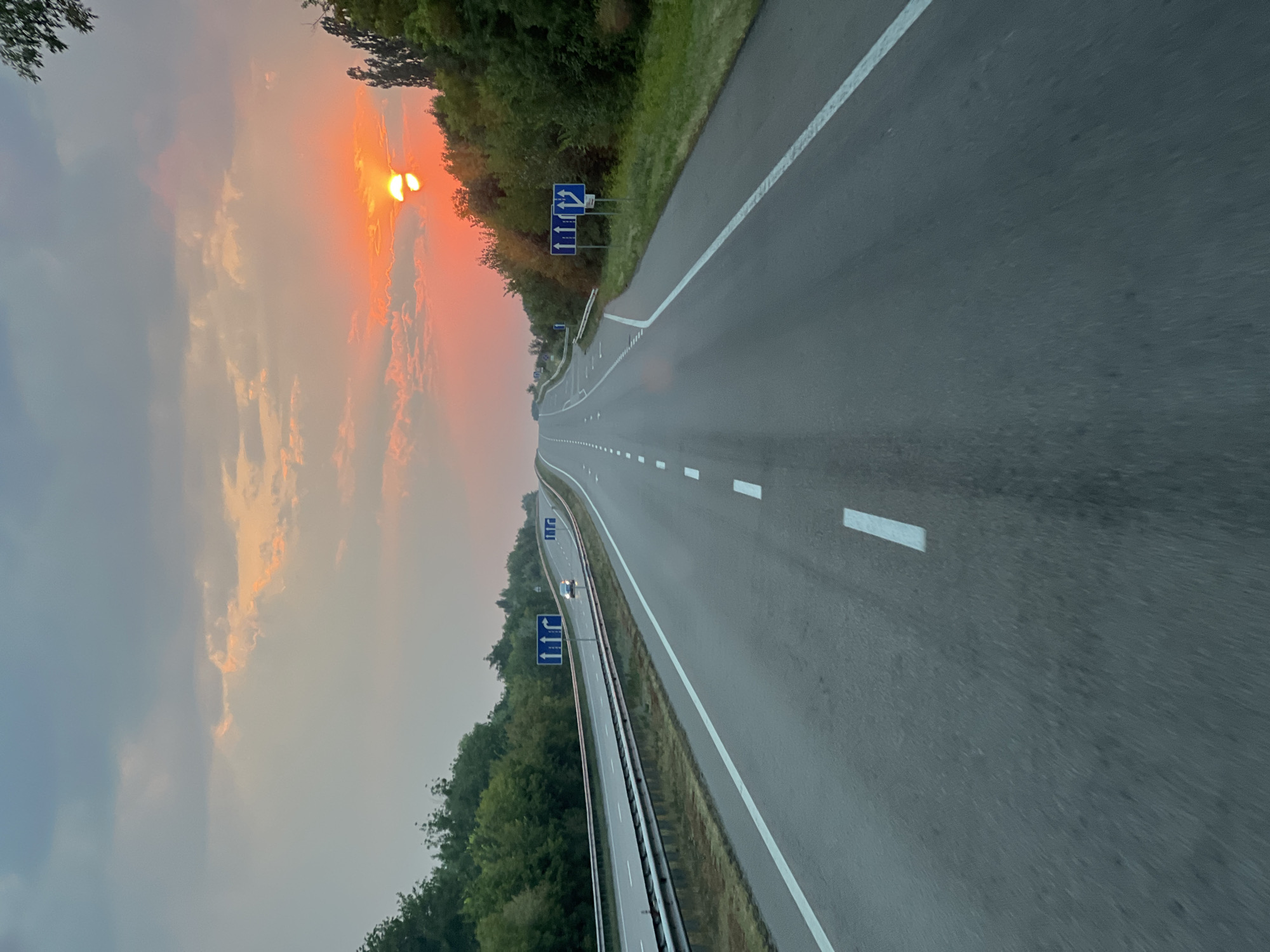 This media file depicts a highway scene at sunset in Nychyporivka, Ukraine. The sun is low in the sky, casting a warm, orange glow through the clouds. The highway stretches out into the distance, with clear lane markings and occasional traffic signs. Trees line the sides of the road, suggesting a rural or semi-rural setting. One sign displays blue directional arrows, indicating multiple lane choices. A single car is visible on the highway, traveling away from the viewer. The overall mood is peaceful and serene, with the setting sun creating a beautiful, tranquil atmosphere. There is no visible text other than the directional signs on the highway.