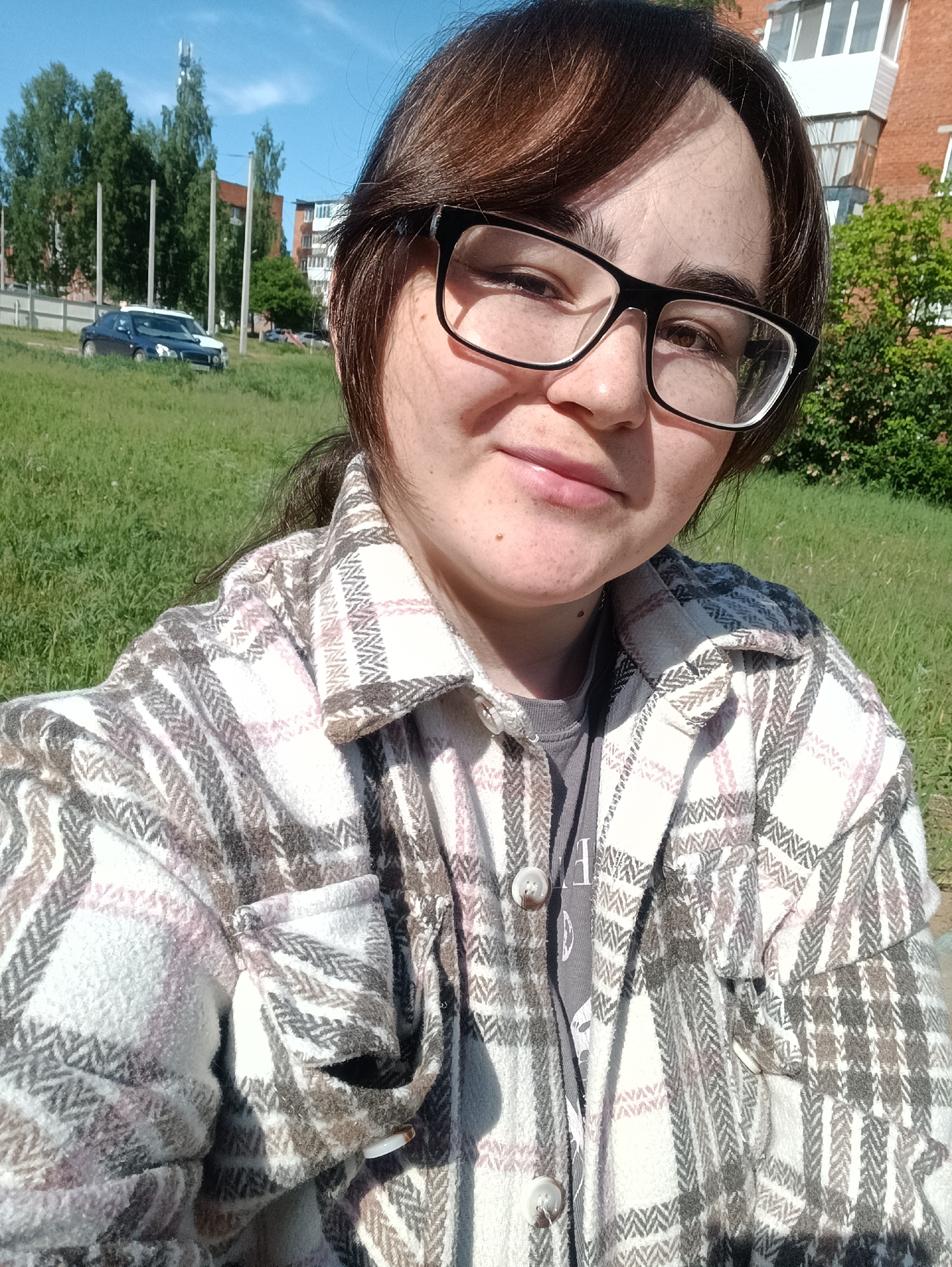 A young woman is captured in a front-facing selfie, smiling slightly and looking directly at the camera. She wears large black-rimmed glasses and has dark hair, styled with bangs across her forehead. Her attire consists of a gray t-shirt, partially visible under a prominent plaid button-up shirt. The plaid pattern features white, light pink/purple, and various shades of brown and gray, with a distinctive herringbone-like texture in some sections. The t-shirt has a partial letter "F" or similar visible on it.
The setting is outdoors on a bright, sunny day, indicated by the clear blue sky and strong sunlight creating shadows on her face and shirt. She appears to be in a grassy area, possibly a park or an undeveloped patch of land near residential buildings. In the background, to her left, two cars are visible parked on or next to a grassy patch, partially obscured by the high grass. One car is dark blue, and the other is white. Beyond the cars, several tall, bare poles stand vertically, and numerous green trees are scattered across the landscape. Further back, an apartment building with red-brown brick facades and white balconies is visible, typical of a residential area in Noviy, Russia, where the image was captured. The overall atmosphere is relaxed and casual.