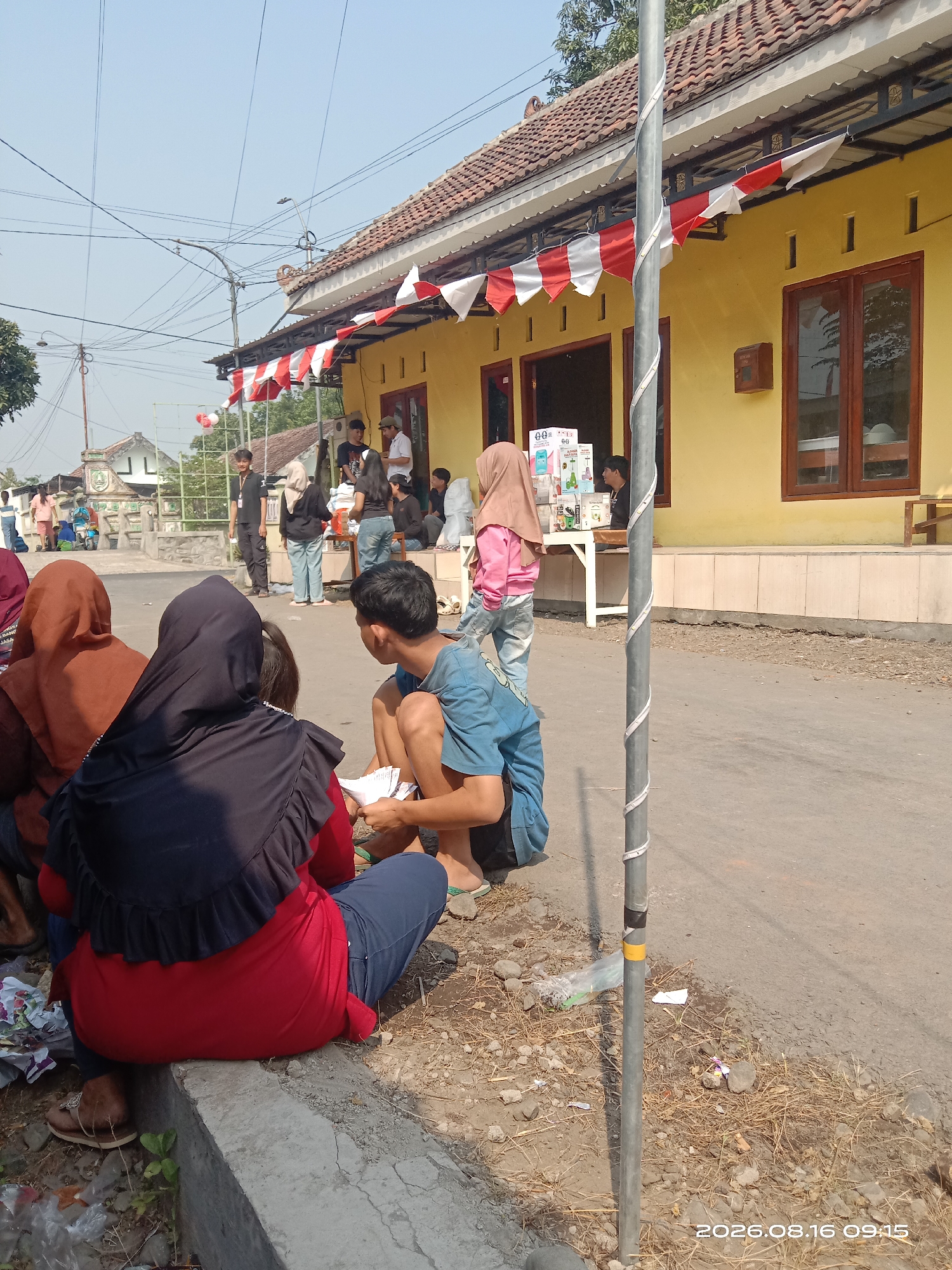 In Banyuarang, Indonesia, on a sunny day, a group of people are gathered outside a yellow building adorned with red and white bunting. The building appears to be a shop or a community center, with wooden-framed windows and a covered entrance. Several individuals are seated on the ground in the foreground, facing away from the camera. One person in a red shirt and black hijab is sitting on a concrete ledge. To their right, a person in a blue t-shirt is seated on the ground, looking at some papers. Further back, a group of people are standing, some interacting with a table that displays several boxes and items, possibly for sale or distribution. In the background, there are trees, a street, and more buildings. A utility pole with a spiral-patterned decoration stands prominently in the middle ground. The overall scene suggests a local gathering or event in a semi-urban or rural setting.