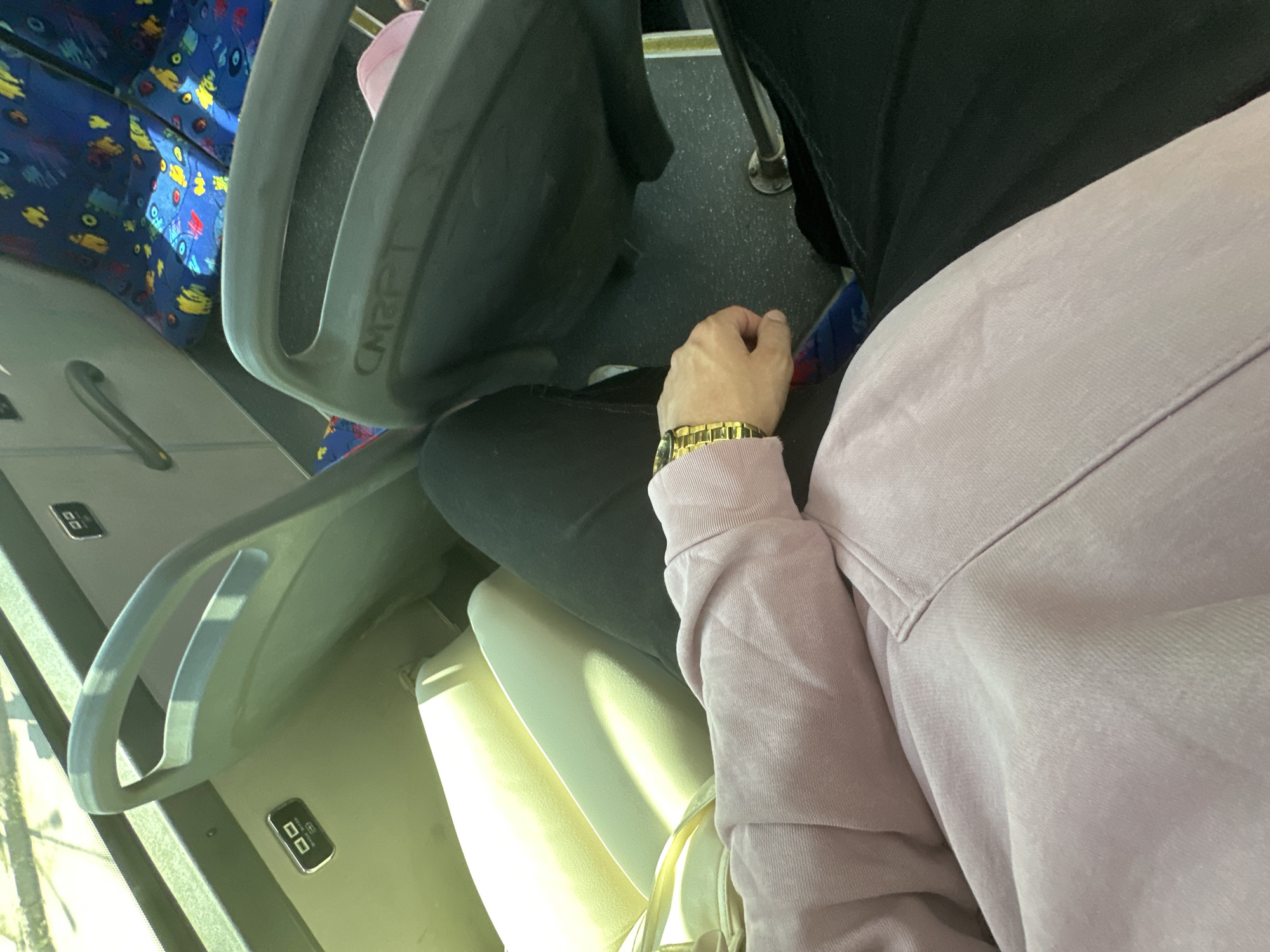An eye-level shot captures a person seated on a bus, with parts of their body and the bus interior visible. The person is wearing a light lavender or pale purple long-sleeved top and black trousers, along with a black head covering or scarf. A prominent golden wristwatch is visible on their left wrist.
In front of the person, there is a light-colored, possibly cream or beige, bus seat. Further down the aisle, other bus seats are visible, upholstered in a vibrant blue fabric with a pattern of abstract yellow, red, and light blue shapes. Sunlight streams in through a window on the left, casting bright highlights on the seats and the person's clothing, suggesting it is daytime with sunny weather.
The bus interior features a dark, textured floor and a grey overhead panel with rectangular black modules that appear to be service buttons or lights, each showing two square symbols. A grey grab handle is also visible overhead. On the side of the seat directly in front of the person, the text "CMRPT" is faintly visible. The scene depicts a quiet moment of travel within the city of Sofia, Bulgaria.