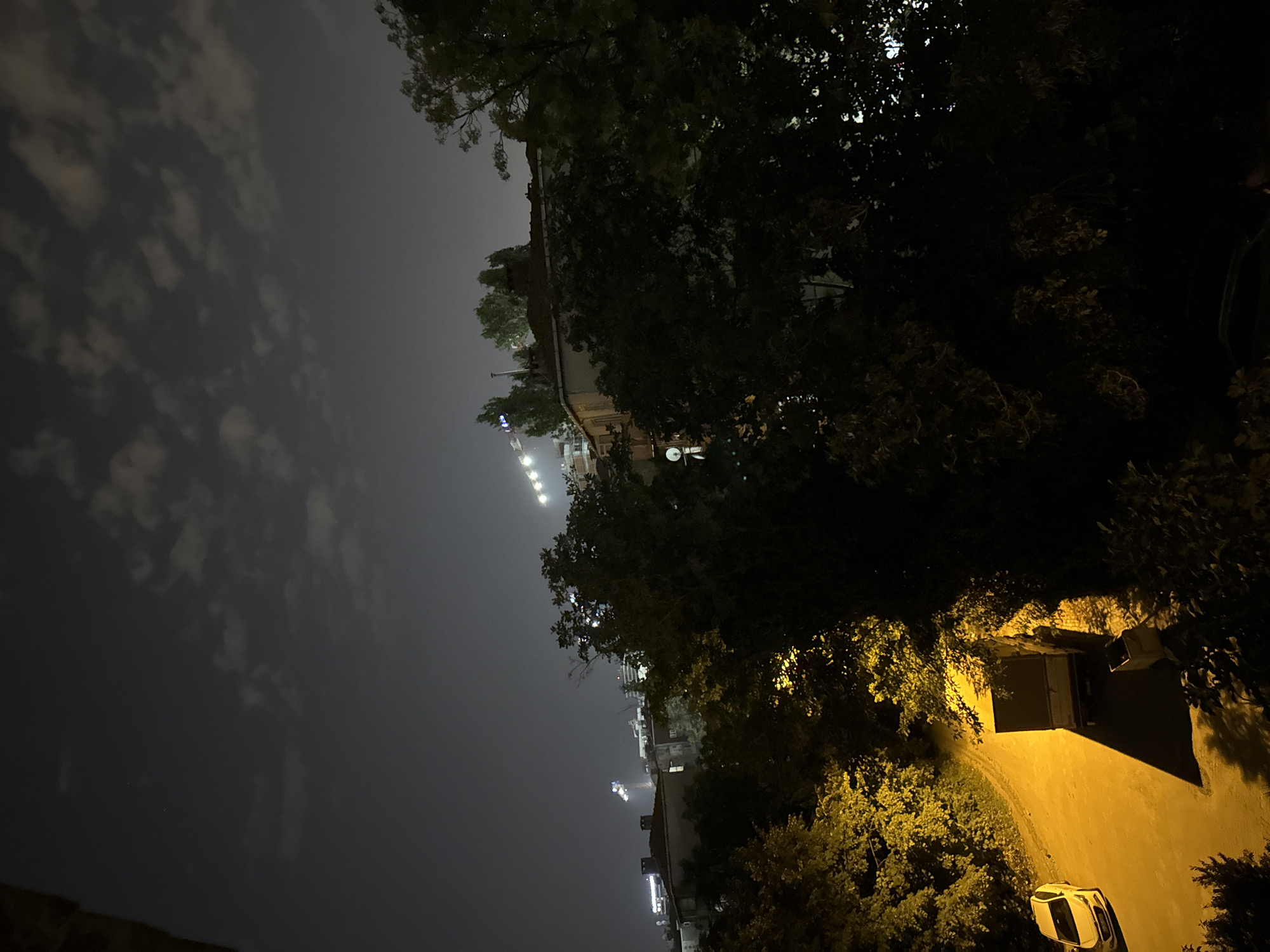 The image shows a nighttime street scene in Gungoren, Turkey. A bright yellow sedan is parked on the left side of a paved road, illuminated by a strong light source. To the right of the car is a large, boxy truck, also parked, casting a long shadow. A trash bin is visible next to the truck. Dense foliage, appearing as dark masses, surrounds the road and the vehicles, with some areas of the trees brightly lit by yellow light, suggesting artificial illumination from streetlights or nearby buildings. In the background, silhouetted buildings and trees are visible against a dark sky with scattered clouds. Distant stadium lights or industrial lighting can be seen on the horizon, creating a hazy glow. The overall impression is one of an urban environment at night.