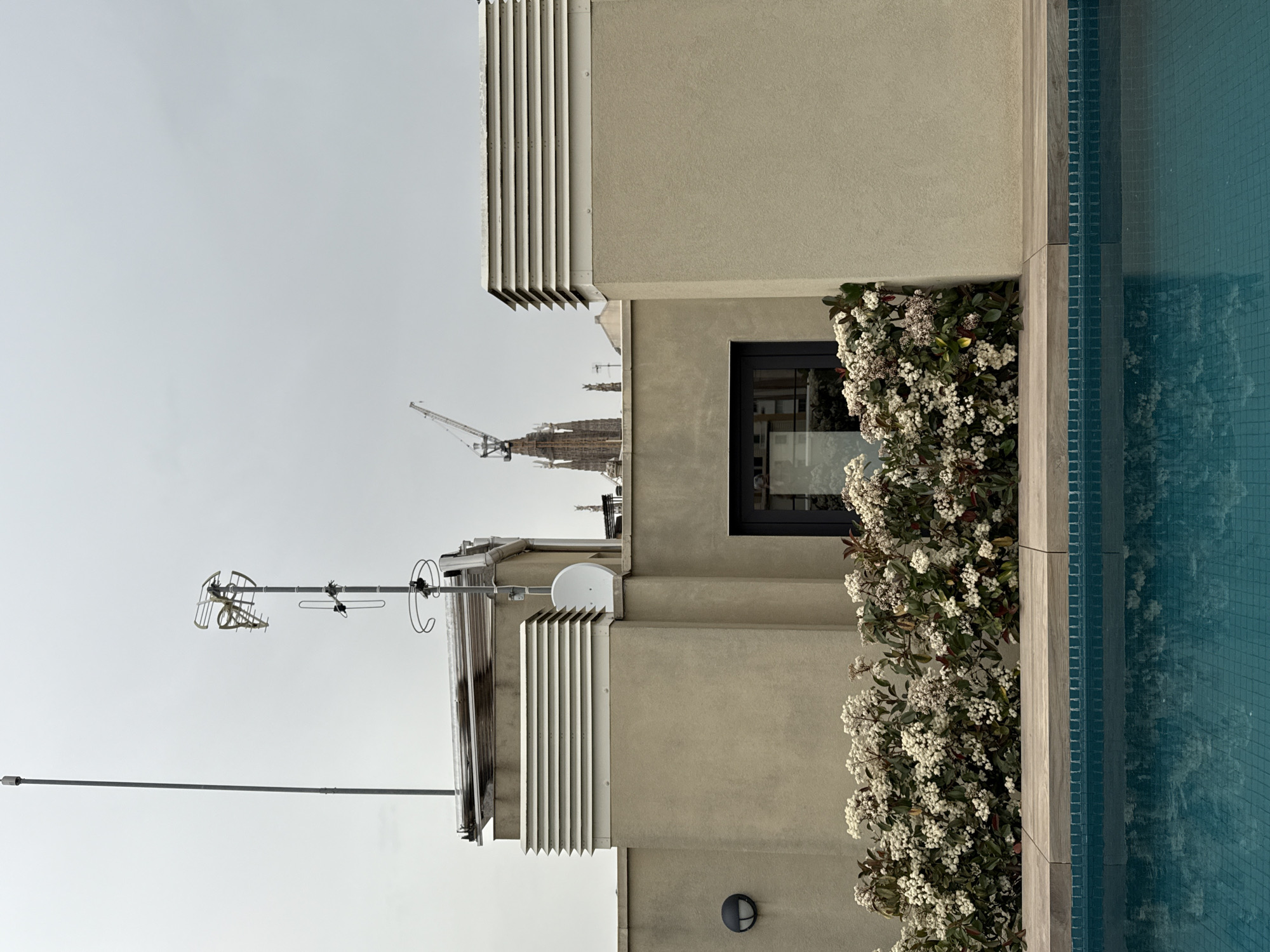 The image depicts the architecture of an urban rooftop in Barcelona, Spain. The scene is characterized by a flat roof featuring a variety of technical installations, including an antenna array and satellite dish, which suggest a residential or commercial building. In the background, the skyline shows the distant silhouette of an unfinished church building, with one or more cranes. The flat roof is adorned with an architectural sunscreen to the building, and there is shrubbery, next to an outdoor pool with turquoise blue water, and the wall of the building.