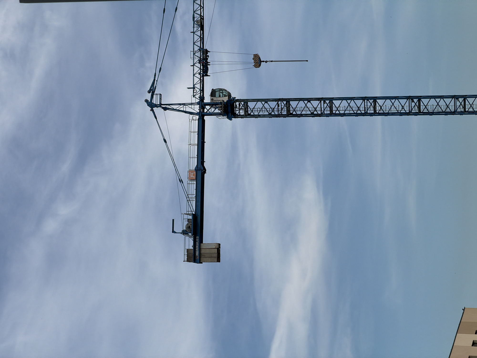 A construction crane is the main subject of the image, silhouetted against a hazy, pale blue sky with wispy clouds. The crane is blue and has a long horizontal boom with a counterweight on the left side, labeled "HERKULES." A cab is visible near the base of the boom. On the right side of the boom, a hook with a yellow and black weight hangs from a chain. In the lower left corner, the top portion of a light-colored building with a dark roofline is visible. The image appears to be taken in Łódź, Poland, as indicated by the presence of a construction crane, which is common in areas undergoing development.
