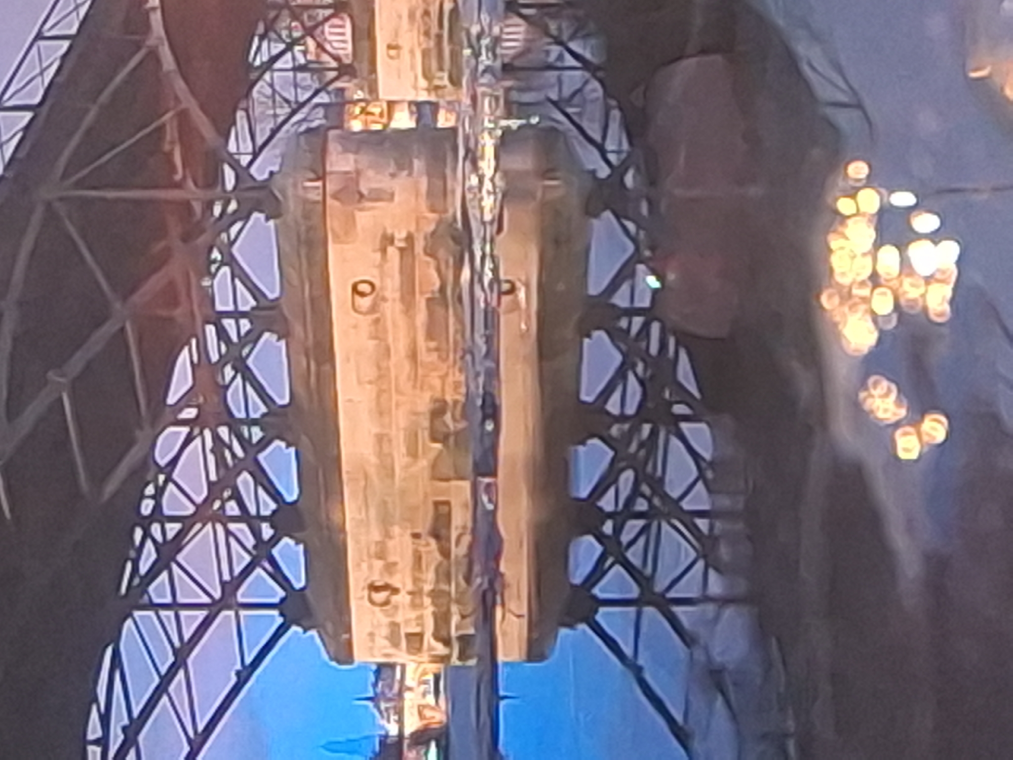 The image shows a stone bridge support in Izmail, Ukraine. The support is made of large, light-colored stone blocks and has two circular openings on its upper surface. It is reflected in the water below, which also reflects the metal structure of the bridge above and some city lights in the distance. The sky appears to be dusk or dawn, with a pale blue hue visible through the bridge's metal framework. There are no people or animals visible in the image.