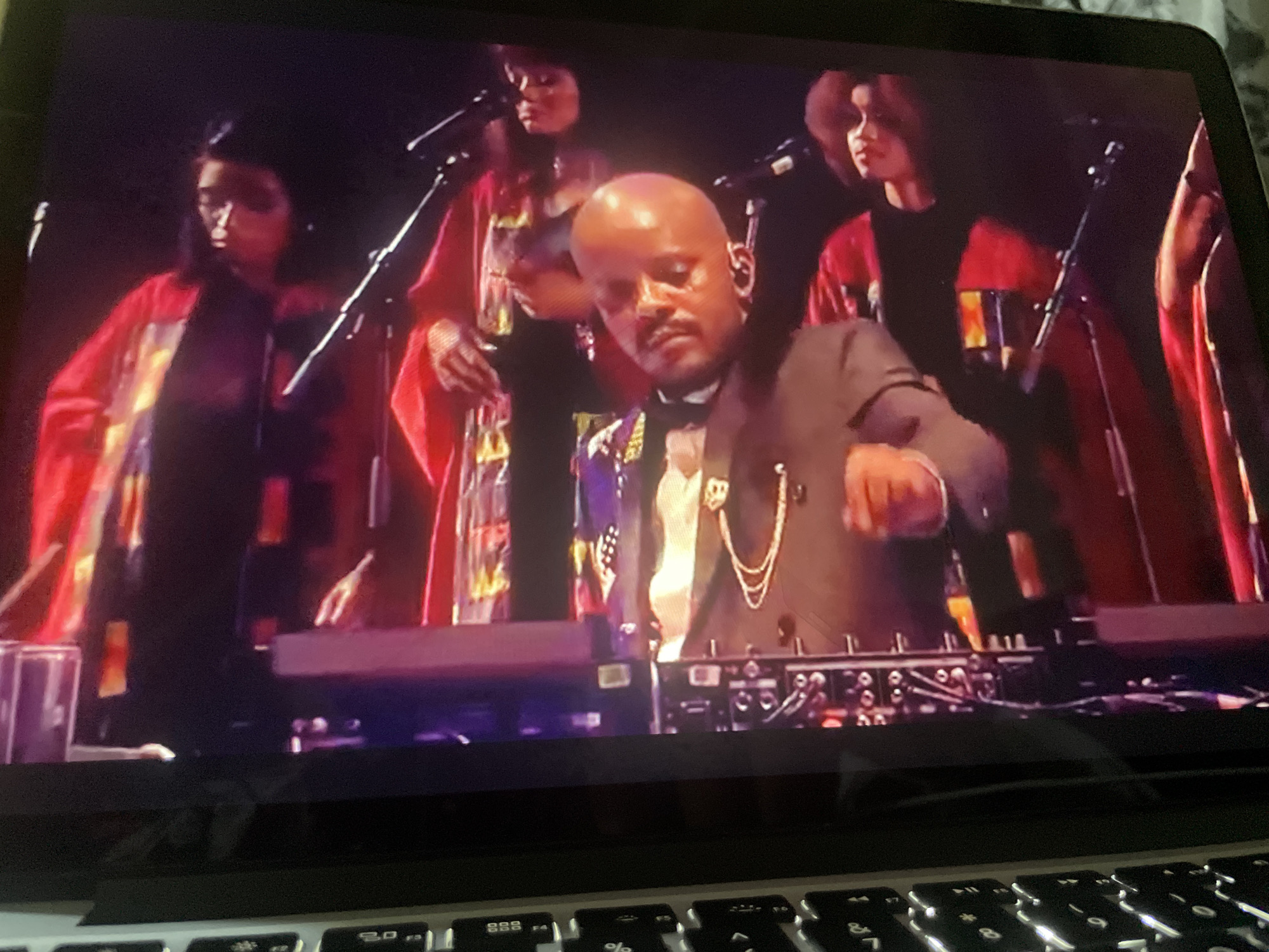 The image is a close-up, slightly angled shot of a laptop screen displaying a live performance. The performance appears to be a musical act with at least three individuals visible on stage. In the foreground of the screen, a bald, dark-skinned man wearing a suit and a gold chain is seated behind what appears to be DJ equipment or a control board. He is looking downwards and his right hand is raised as if operating the equipment. Behind him and to his sides, two women in matching red, hooded robes are standing, each with a microphone in front of them. The lighting on stage is dim with spotlights highlighting the performers. The laptop is open, and the keyboard is visible at the bottom of the image, out of focus. The scene is indoors, likely a concert venue or studio.