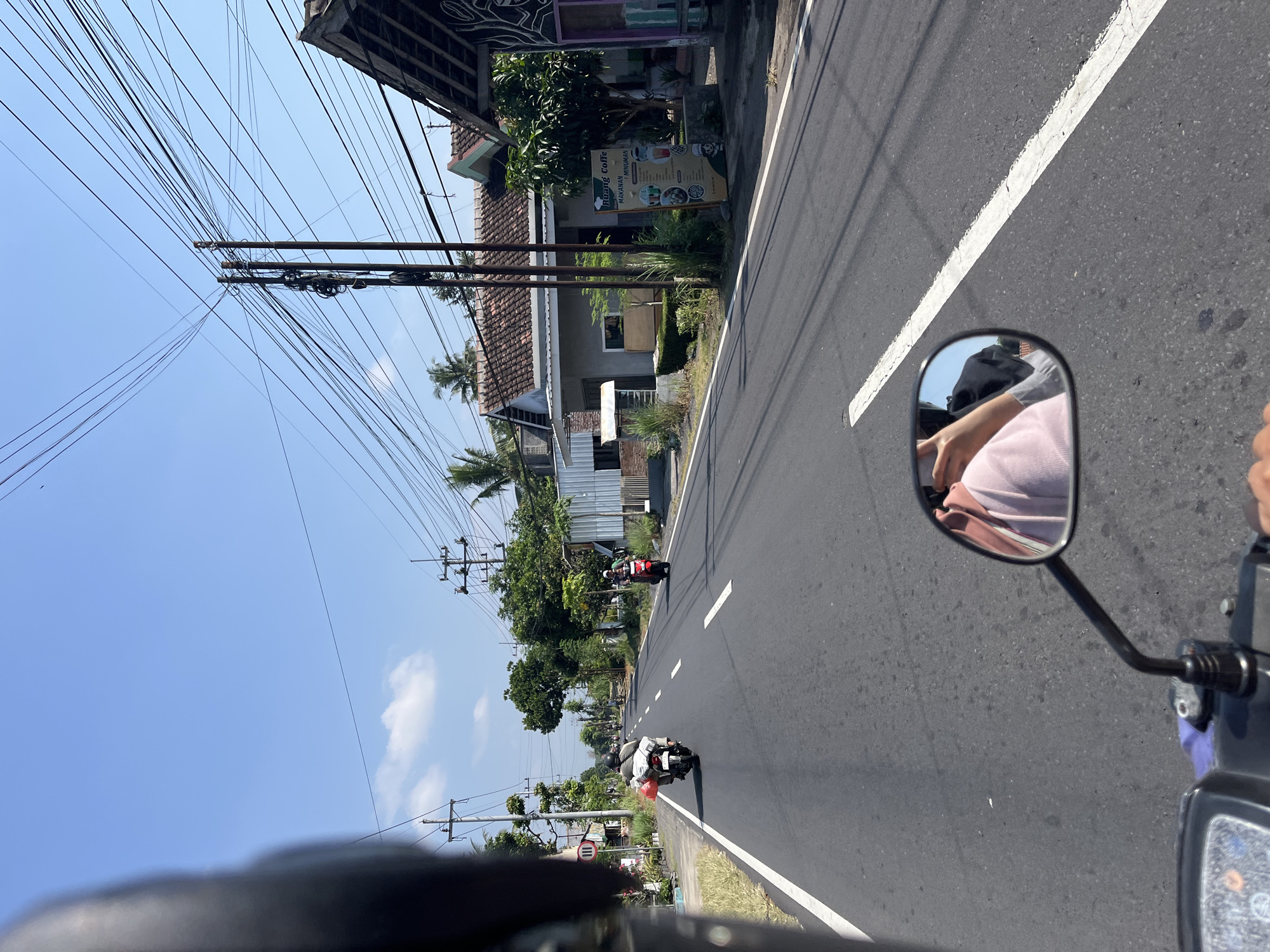 The image depicts a view from the perspective of someone riding a motorcycle on a paved road in Purworejo, Indonesia. The road is lined with buildings and vegetation, with power lines overhead. In the foreground, a motorcycle's handlebar and side mirror are visible. The side mirror reflects the rider wearing a pink hoodie and the back of a person in a grey shirt. Two other motorcycles are seen ahead on the road; one is carrying a red and white package, and another is red. A sign for "HUANG COFFE MAKANAN & MINUMAN" is displayed on the right side of the road. The sky is clear and blue.
