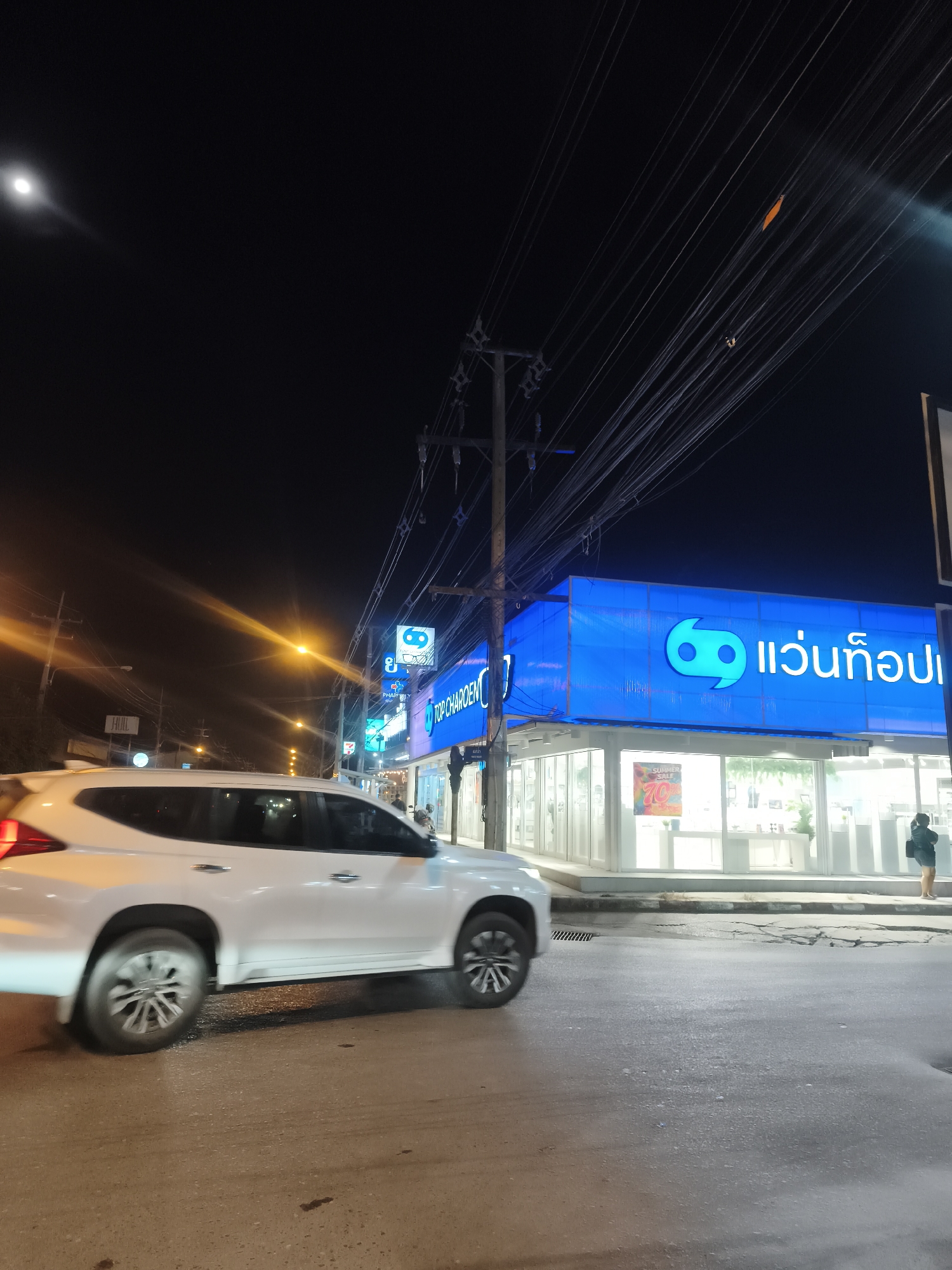 This media file captures a vibrant night scene on a commercial street in Baan Mae Nam, Thailand. The primary focus is a brightly lit storefront of an optical shop, "แว่นท็อปเจริญ" (Wan Top Charoen), identifiable by its large blue sign with a distinctive "69" or abstract logo and English text "TOP CHAROEN."
A white SUV is prominently featured in the foreground, traveling from left to right. Its wheels are blurred, indicating motion. The road surface appears damp and reflective, casting shimmering light patterns. On the sidewalk in front of the optical store, a single person is visible, standing near the building's brightly illuminated windows, where a "SUMMER SALE 70%" poster is displayed.
The sky is dark, characteristic of nighttime. A very bright light source, likely a distant streetlight or the moon, is visible in the upper left, emitting a distinct lens flare. The street is lined with multiple power poles from which an intricate and dense network of overhead electrical wires extends across the frame. Another power pole is visible to the left, and numerous streetlights cast warm yellow and orange glows further down the road, creating long light streaks due to lens flare.
Additional visible text includes "PHARMACY" and "7-Eleven" signs further along the street, indicating other businesses in the vicinity. A sign for "HUILL" is also partially visible on the far left. The overall setting is a bustling urban or suburban area at night, characterized by artificial light, traffic, and commercial activity.