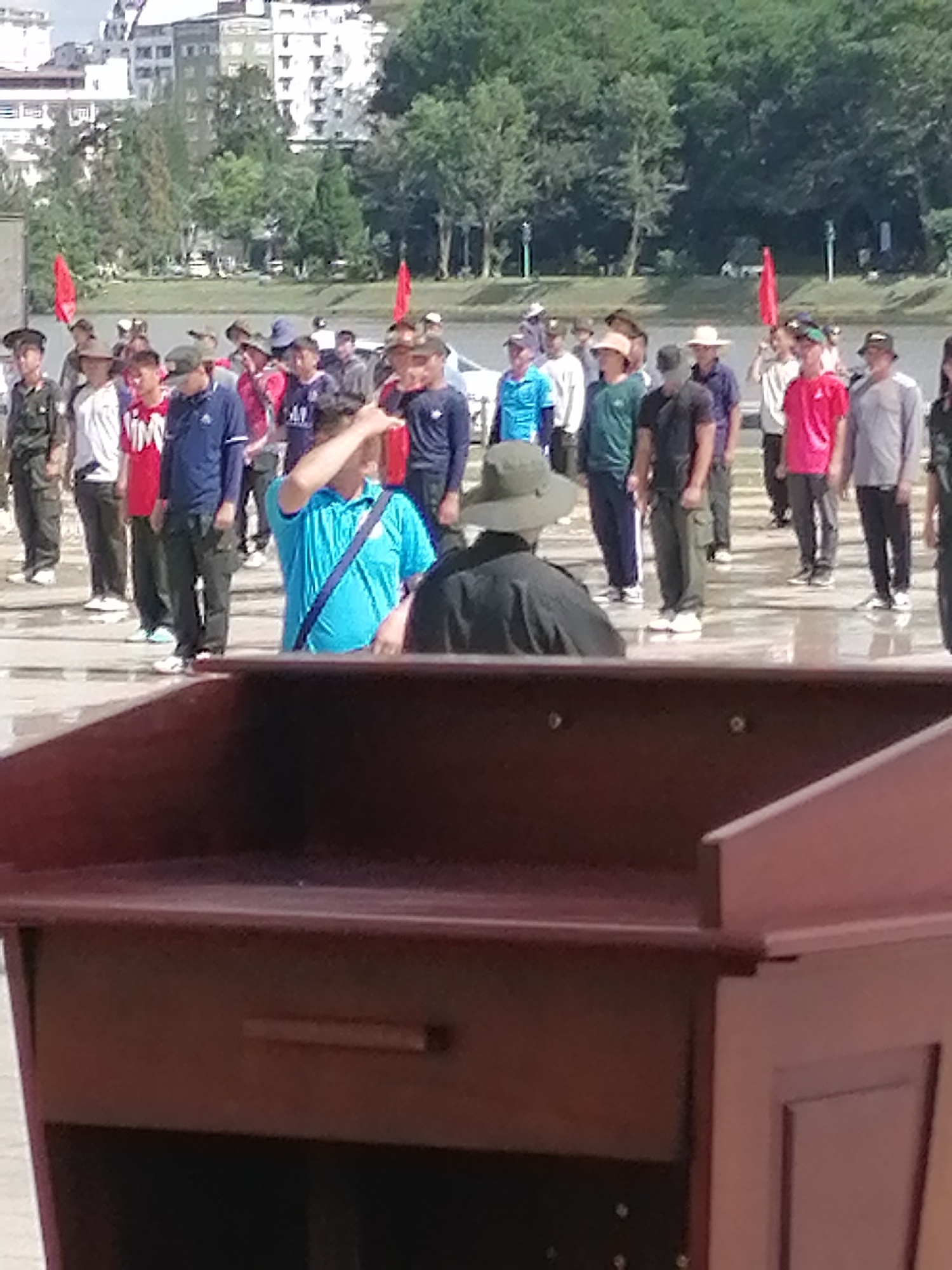 In the foreground, a dark reddish-brown wooden podium with drawers is visible, obscuring the lower half of the scene. Behind it, a group of people are standing in formation outdoors. Most are wearing casual clothing such as t-shirts, shirts, and pants, with some in what appear to be military-style uniforms. One man in the middle is wearing a bright blue shirt and a dark blue bag slung across his shoulder, with his right hand raised to his forehead. Another individual to his left is wearing a khaki-colored hat and a dark shirt. A person wearing a bright red t-shirt is standing in the background among the crowd. Red flags are visible in the distance, planted in the ground. The background shows lush green trees and buildings, suggesting an urban setting. The ground appears wet, possibly from rain or a water feature. This scene likely depicts a public gathering or event near Xuan Huong Lake in Da Lat, Vietnam, given the combination of outdoor space, trees, buildings, and the general atmosphere often associated with such locations in the city.