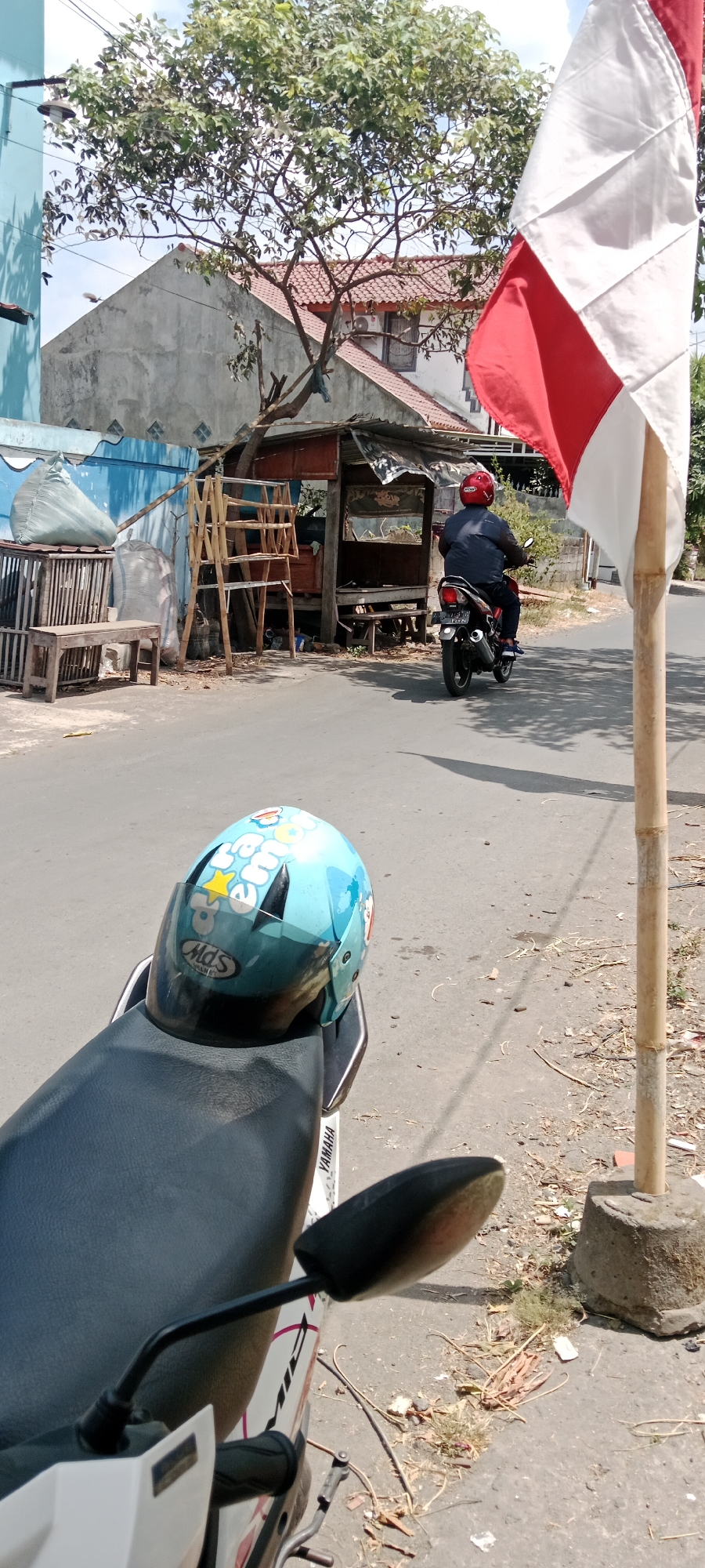 In the city of Purwomartani, Indonesia, a motorcycle is parked on the side of a paved road. A light blue helmet with cartoon characters is placed on the seat of the motorcycle. The helmet has a clear visor and the brand "MDS" is visible on it. The motorcycle has a "YAMAHA" logo on its side panel.
In the background, a person wearing a red helmet and a dark jacket is riding a black motorcycle away from the viewer. The motorcycle has a license plate that is partially visible. Next to the road, there is a small, weathered wooden stall with a corrugated metal roof. Various items are stacked around the stall, including a wooden bench, a wooden rack, and large sacks. A portion of the Indonesian flag, red and white, is visible on a bamboo pole to the right of the frame. The environment appears to be a street scene with some vegetation and buildings in the background, under a bright, sunny sky. The ground is paved with asphalt and there is dry grass and debris along the side of the road.