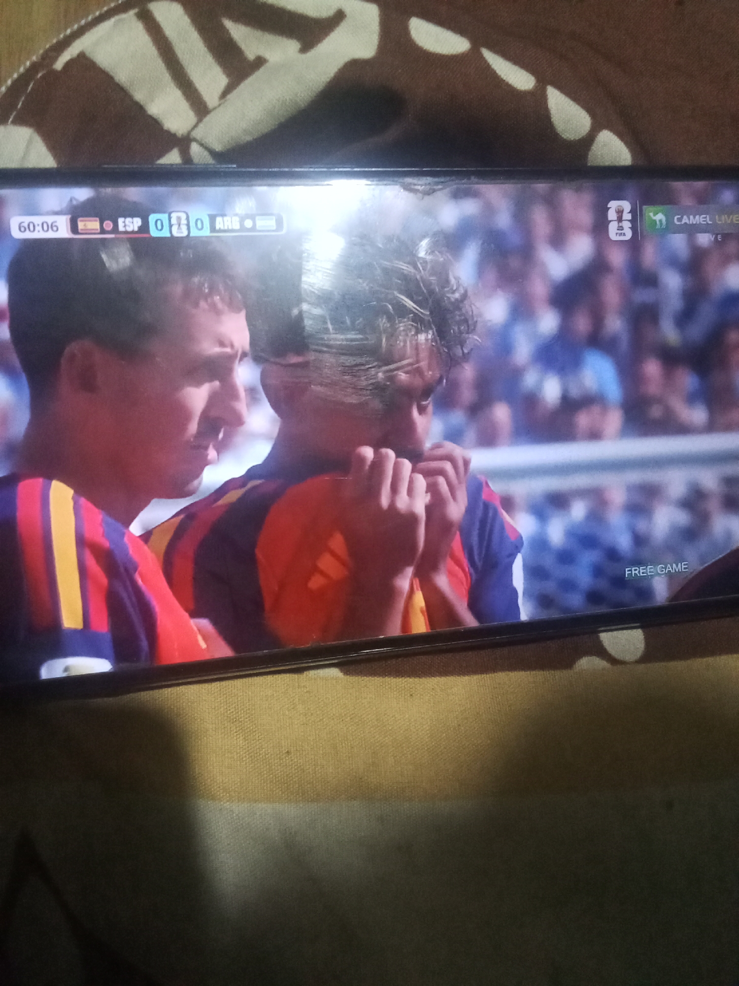 The image shows a television screen displaying a soccer match. The score is 0-0 between ESP and ARG at 60:06. There are two soccer players visible on the screen. The player on the left is looking forward with his mouth slightly open. The player on the right is touching his mouth with his hands. The background shows a crowd of spectators. In the upper right corner of the screen, there is a FIFA logo and text that reads "CAMEL LIVE". In the lower right corner, it reads "FREE GAME". The television is placed in front of a patterned fabric that appears to be a blanket or tablecloth. The scene is dimly lit. The location of Buketglumpang, Indonesia is not discernible from the image.