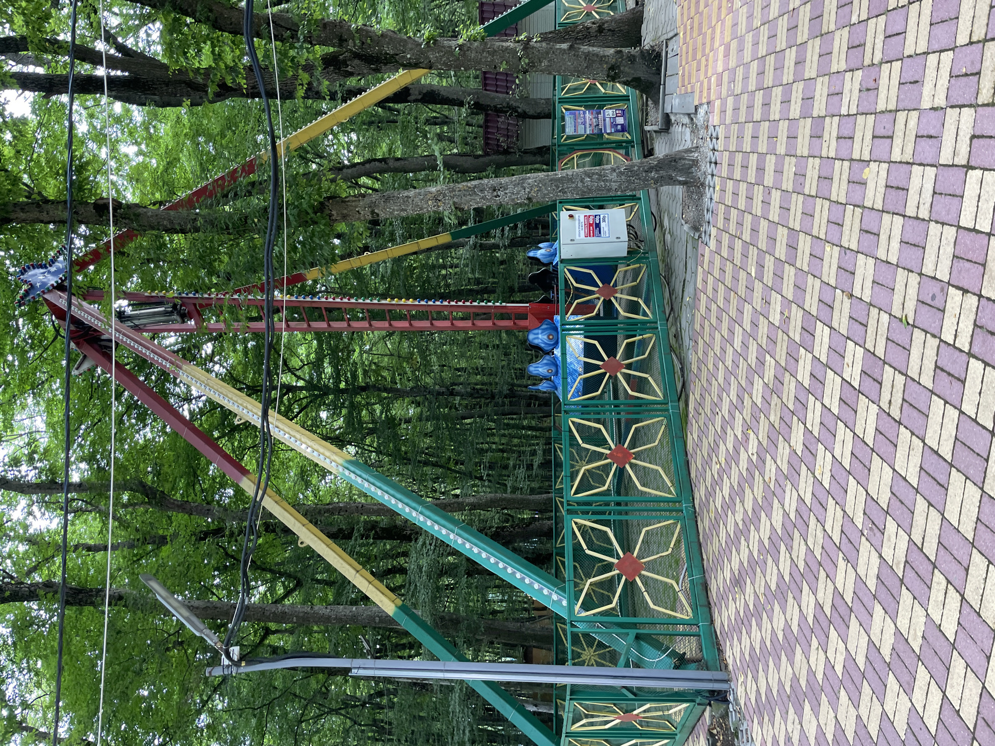 The image depicts an amusement park ride with a large pendulum-like structure featuring colorful arms (red, yellow, and green) and lights, set against a backdrop of dense green trees. In the foreground, a decorative green metal fence with yellow and red accents surrounds the ride's operational area. Blue seats are visible on the ride's gondola. A paved pathway with a diamond pattern of purple and beige tiles leads to the ride. A white electrical box with Cyrillic text is mounted on the fence. The environment appears to be a park or outdoor recreational area. The ride is located in Stavropol, Russia.