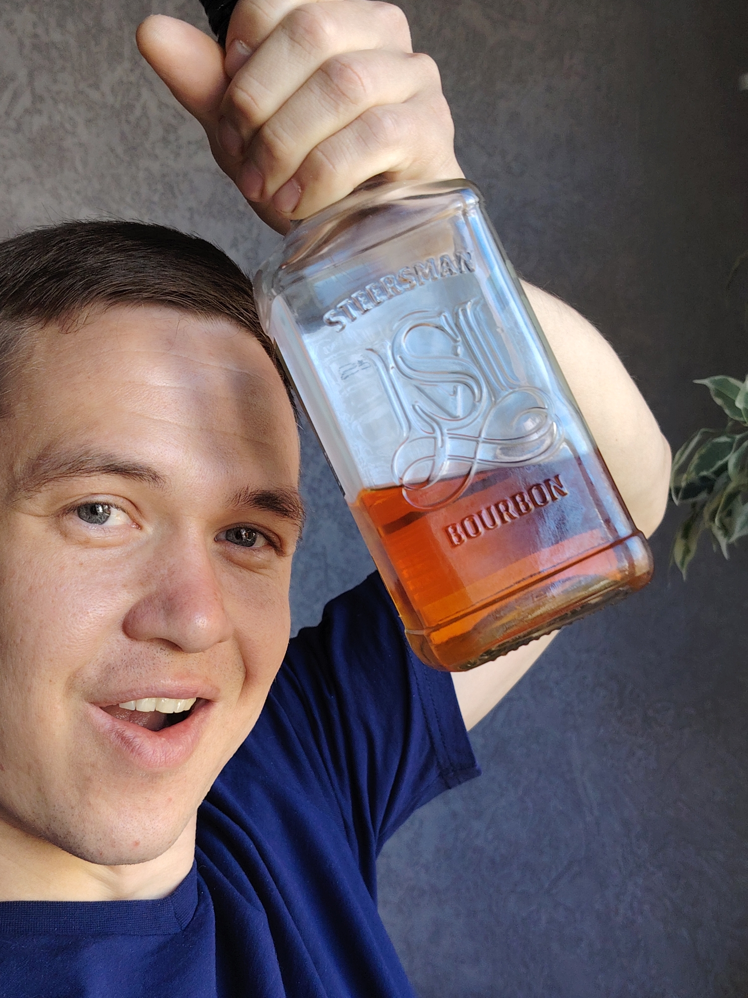 A man is holding a bottle of bourbon in front of him. The bottle is labeled "Steersman Bourbon." The man is wearing a blue t-shirt. The background appears to be a wall, and a plant is visible to the right of the man. It is impossible to determine the location in the city of Chaikovsky, Russia, from the image.