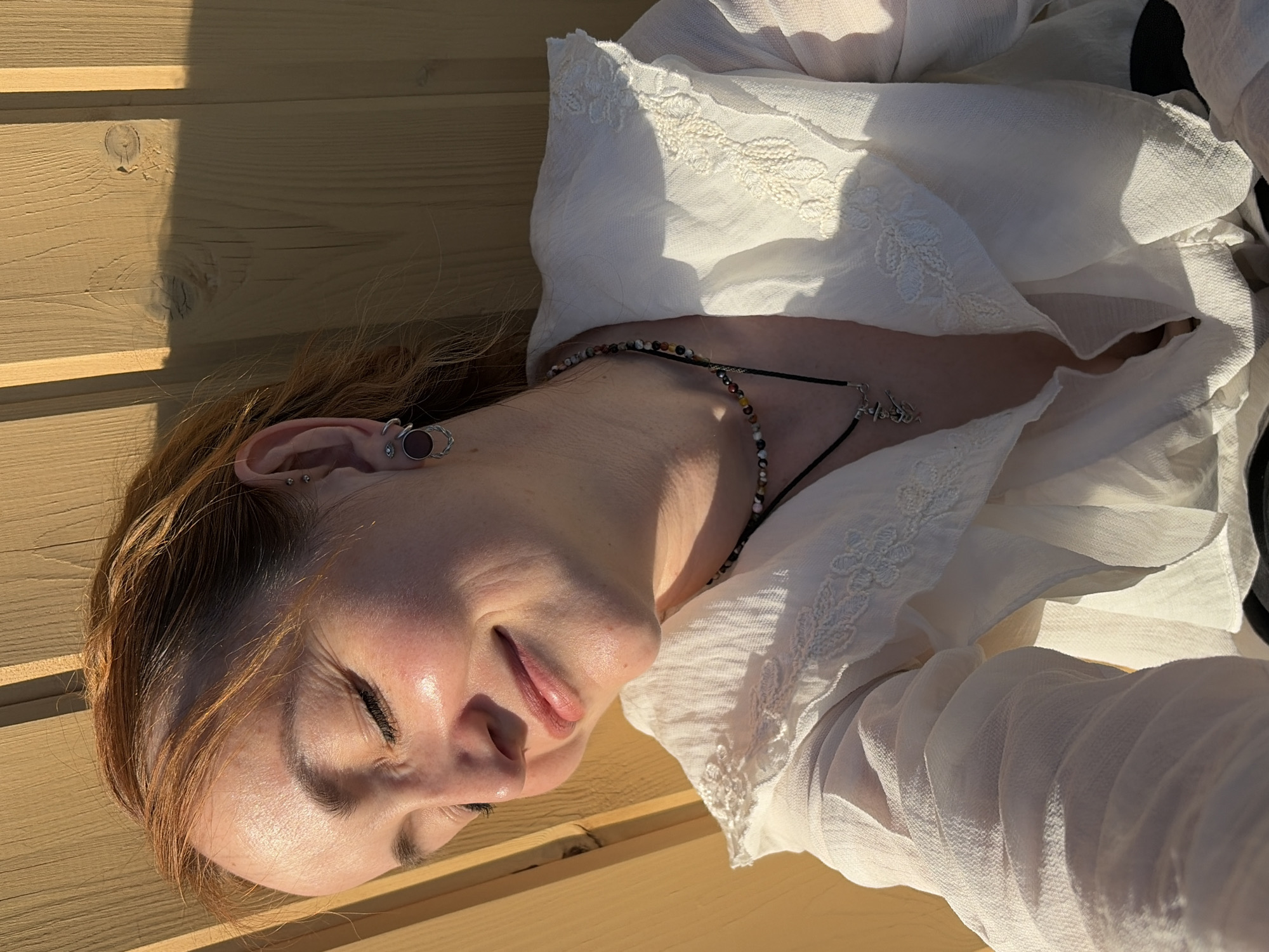 The image shows a light-skinned person with reddish-brown hair lying on a wooden surface, possibly a bench or deck. The person is wearing a white, ruffled blouse and a necklace with a silver pendant. The person's eyes are closed, and they appear to be smiling slightly. The background consists of wooden planks, and there are shadows cast across the surface. The location is unknown as there are no features to determine that location is Chaikovsky, Russia.