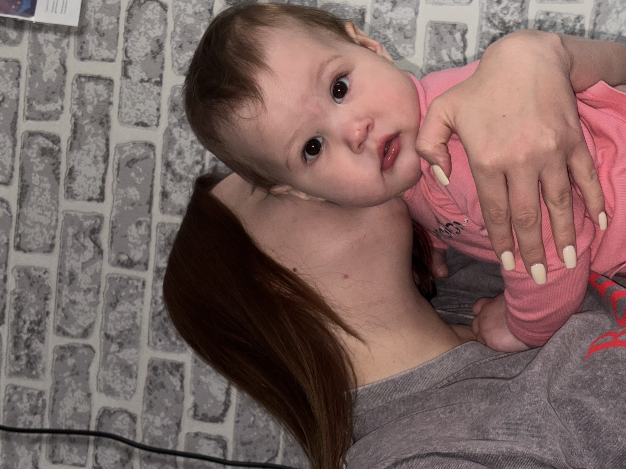 A baby in a pink shirt rests against a person's shoulder. The person has long, reddish-brown hair and wears a gray shirt. The person's hand, with light-colored manicured nails, is positioned around the baby. The background appears to be a wall with a faux-brick pattern. It is not possible to determine the specific location within Novopokrovskaya, Russia, from this image.