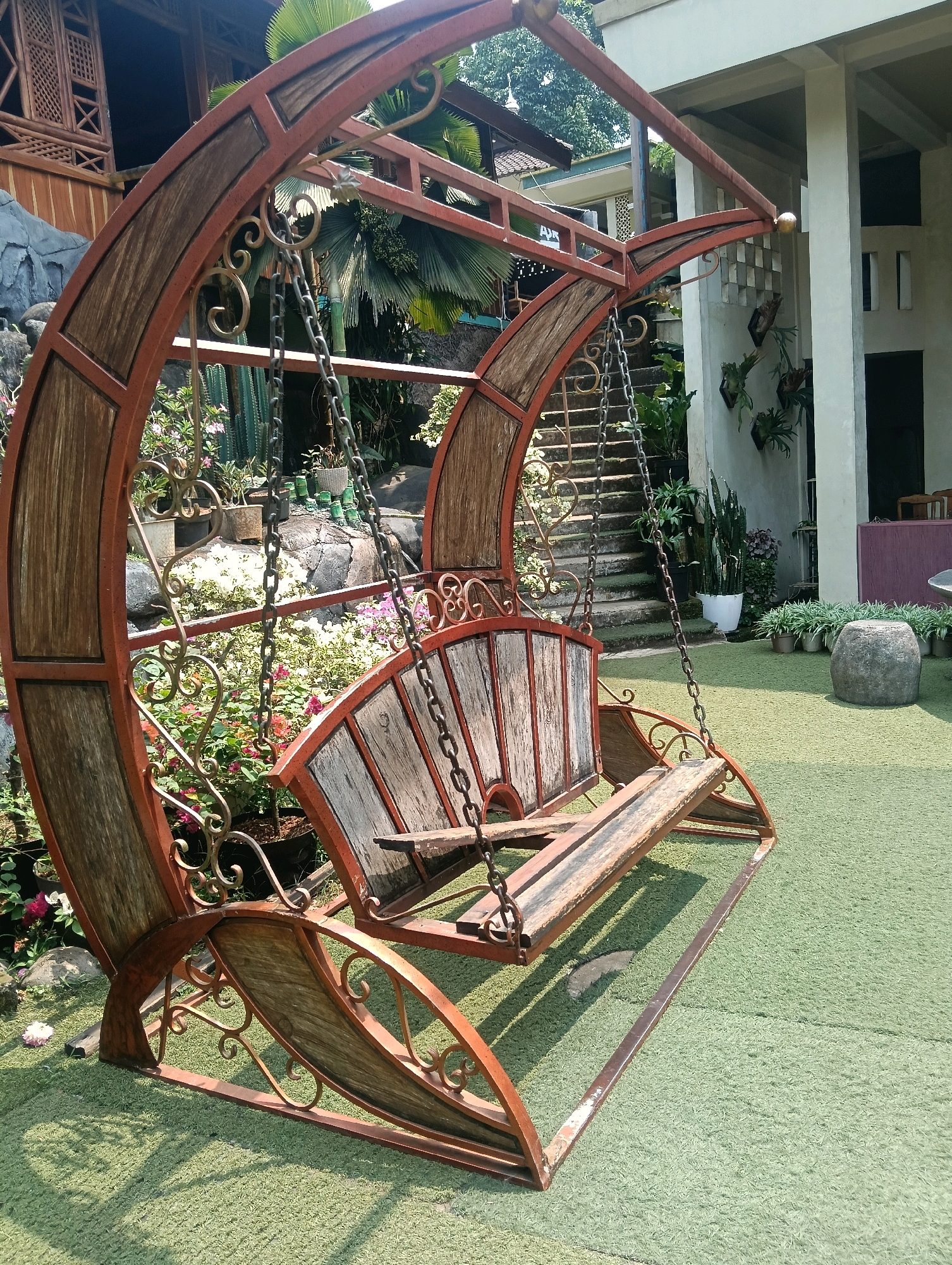 The image captures a rustic, ornate garden swing with a curved metal frame and wooden planks, set on a patch of artificial turf. The swing is designed with decorative metal scrollwork and appears to be empty. Behind the swing, a stone pathway leads up a set of outdoor stairs, flanked by lush greenery and various potted plants. In the background, a multi-story building with white pillars and wooden architectural elements is visible, suggesting a resort or residential area. To the left of the swing, a rocky landscape with small flowering plants adds to the garden aesthetic. The scene is illuminated by natural daylight, casting shadows across the turf. The location is identified as Sempu, Indonesia.