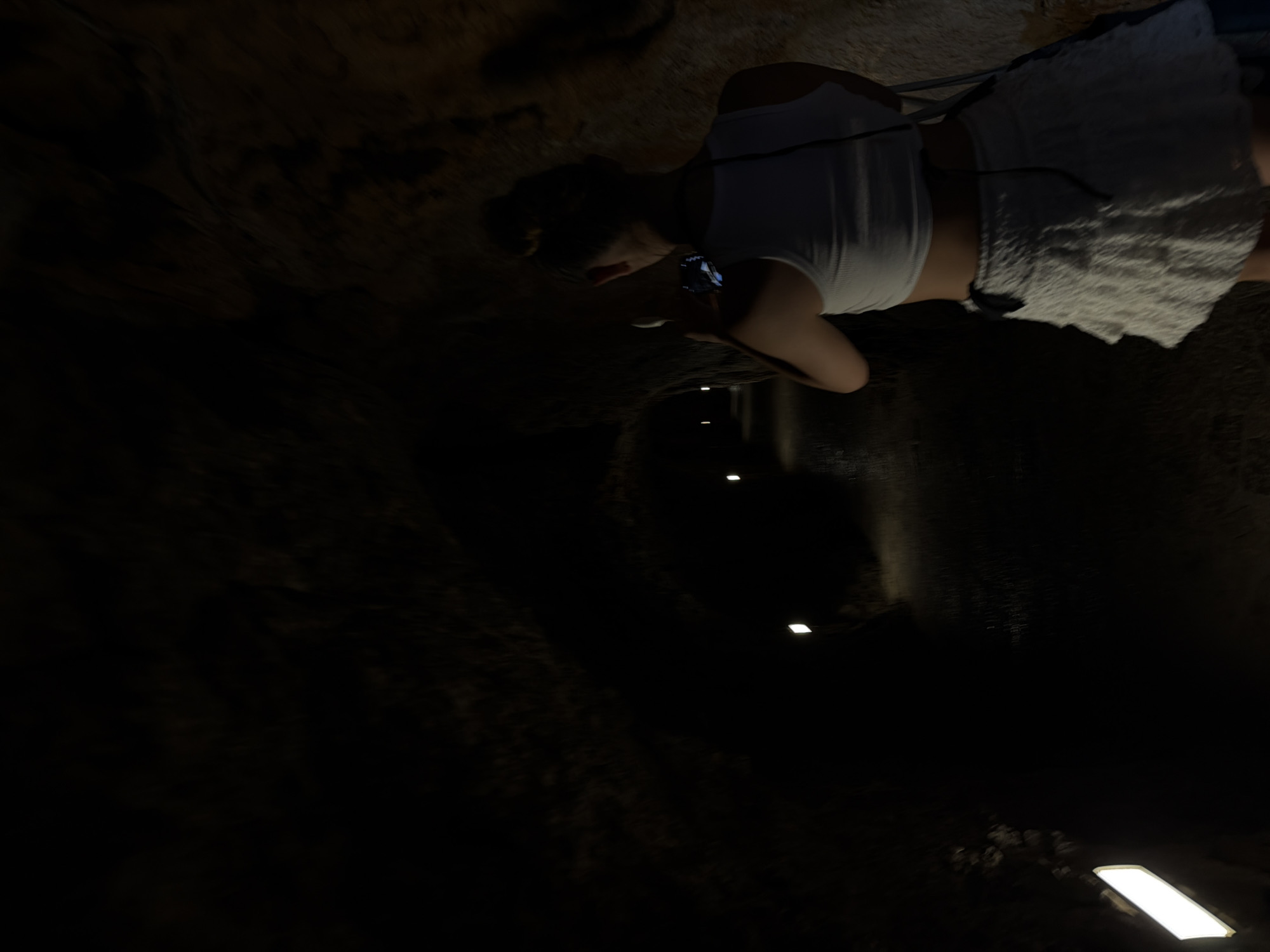The image captures a scene within the Cuevas de Campanet caves, located in Escorca, Spain. The focus is on a woman in the upper right of the frame who is looking into the cave. She is wearing a white tank top and white shorts. The cave itself is dark, with lighting fixtures illuminating the path. The walls are textured with natural rock formations typical of caves.