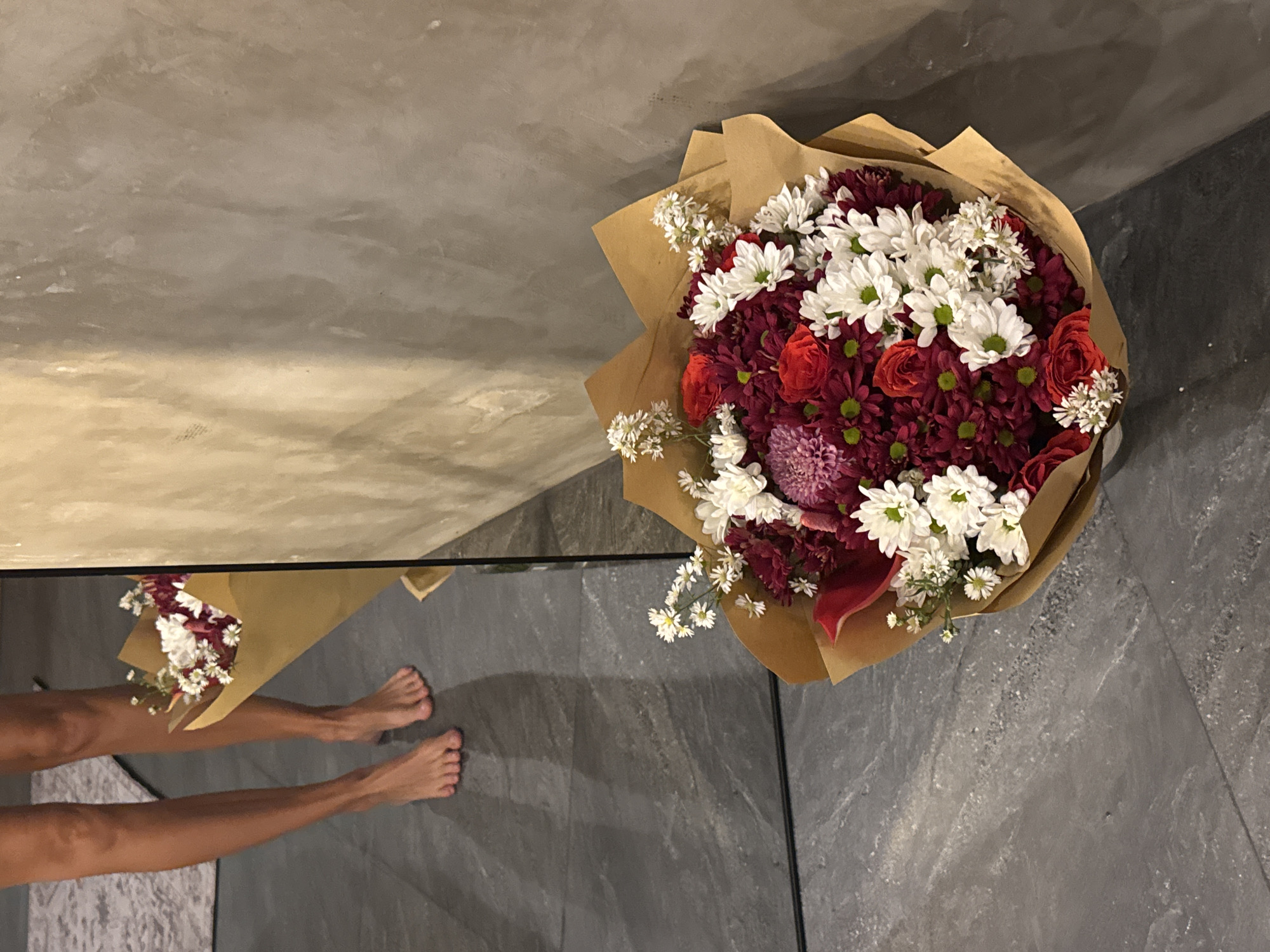 The scene, likely captured in North Kuta, Indonesia, presents a bouquet of flowers (mainly white, red, and burgundy varieties, wrapped in brown paper) resting on a dark gray tiled floor. Reflected in the polished floor, which acts as a mirror, are a second, identical, bouquet and bare female legs and feet. The background of the reflected portion shows a wall painted in shades of beige, while the wall above the tiled floor is painted in a matching dark gray.