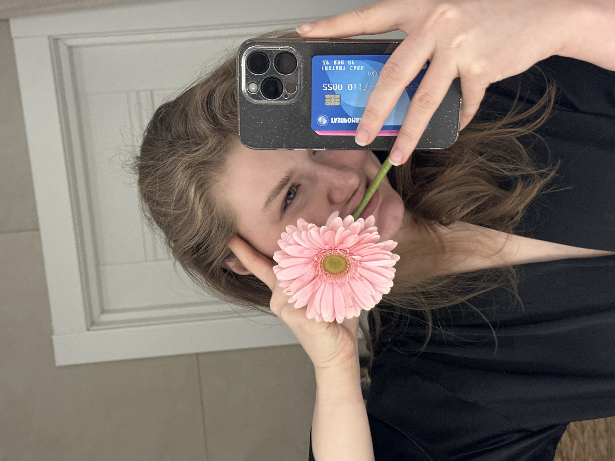 The image is a mirror selfie depicting a young adult female indoors. She has light brown hair and is wearing a black garment. Her left hand holds a pink Gerbera daisy, with its stem positioned near her mouth, partially obscuring her face and revealing a subtle smile. Her right hand holds a dark-colored smartphone, featuring a triple-camera module and a speckled case. A blue credit card is attached to the back of the phone, displaying partial numbers "5500 0173" and reversed Cyrillic text. The background consists of a plain, light-colored wall with subtle horizontal lines and a white paneled interior door. The specific location within Kazan, Russia, is not discernible from the visual evidence.