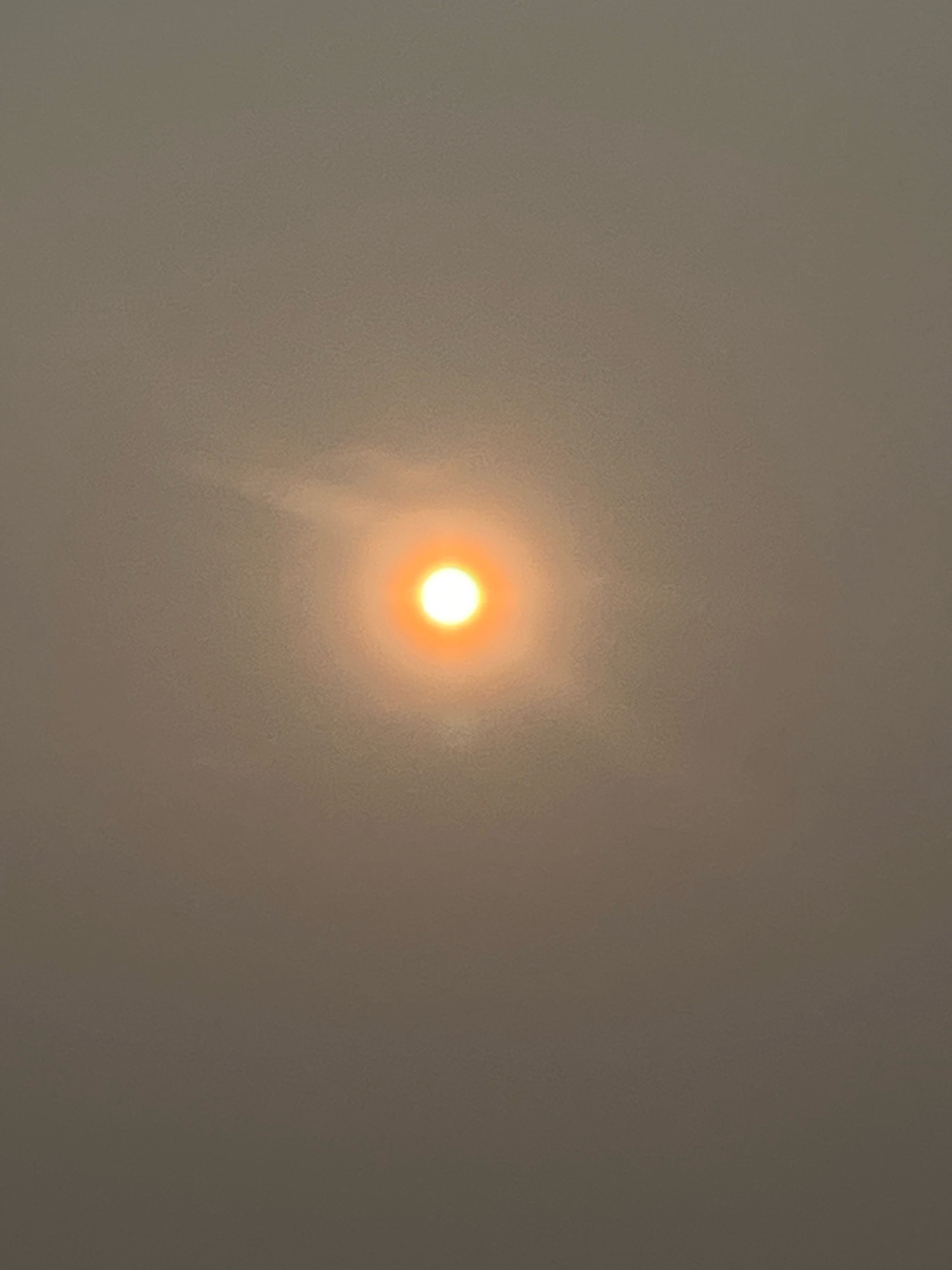 A photograph taken in Kuala Lumpur, Malaysia shows the sun setting through a haze of smoke. The sun appears as a bright orange disk in the center of the image, surrounded by a soft glow. The sky is a uniform hazy gray, obscuring any details of the surrounding environment. The overall impression is one of muted light and subdued color.