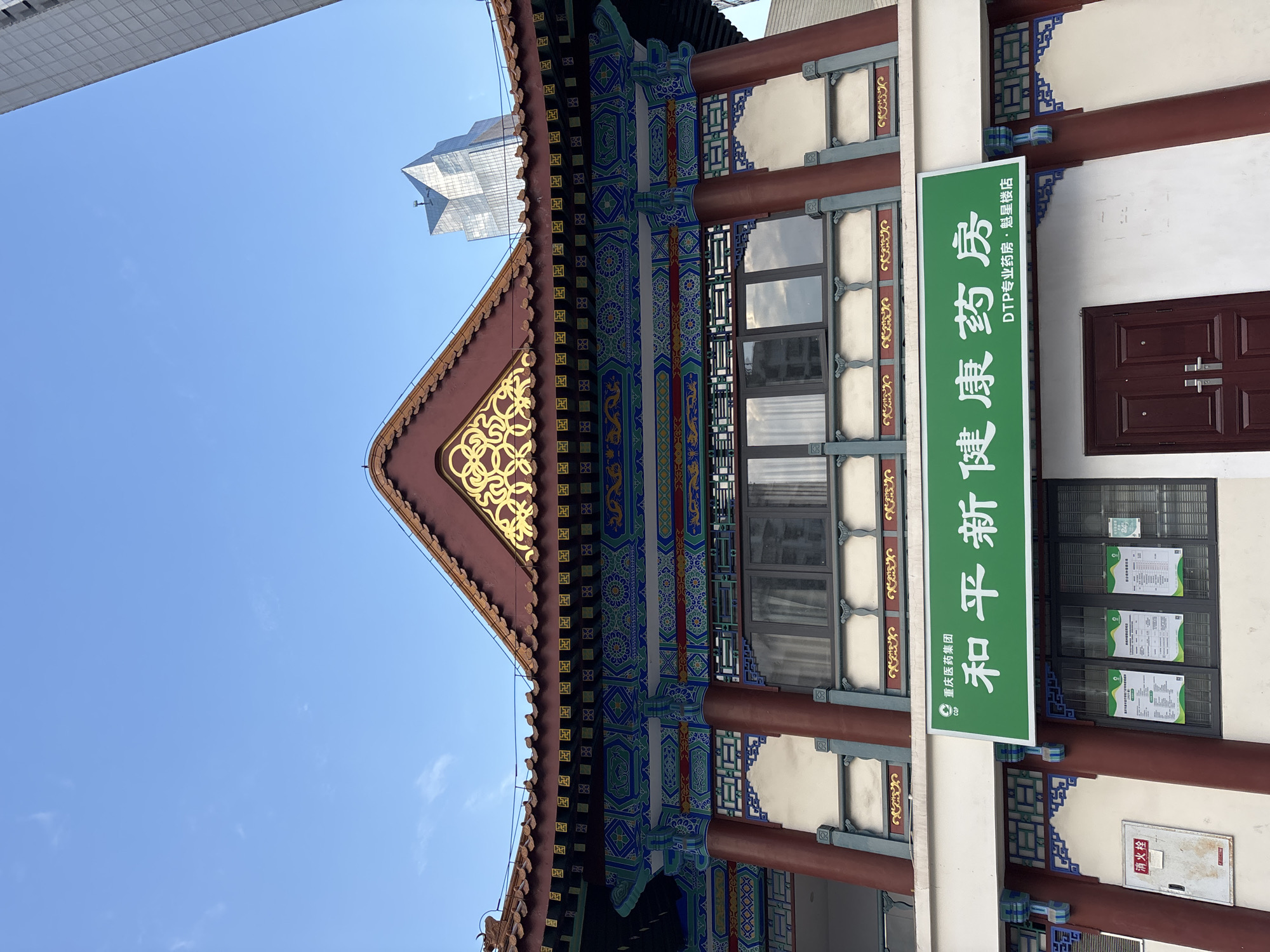 A low-angle medium shot captures the ornate facade of a traditional Chinese-style building, likely a pharmacy, set against a clear blue sky with a few wispy clouds. The building's roof features intricate red and gold detailing, with ceramic tiles and decorative elements. The eaves are adorned with colorful paintings and geometric patterns in shades of blue, green, and red. Below the eaves, large windows reflect the sky and surrounding structures, including a modern glass skyscraper in the background.
A prominent green sign with white Chinese characters is mounted above the entrance, reading "和平新康药房" (Hépíng Xīn Kāng Yàofáng), which translates to "Heping New Health Pharmacy." Below this, in smaller text, it says "DTP专业药房·慰星楼店" (DTP Zhuānyè Yàofáng · Wèixīng Lóu Diàn), indicating it's a DTP professional pharmacy, specifically the "Weixing Building Store." The building's lower level has white walls, red columns, and decorative blue and gold latticework.
To the left, a red metal fire alarm box is mounted on the wall. Several posters are displayed behind glass panes in a window to the right of the fire alarm. A pair of dark red double doors with silver handles is visible at the bottom center of the frame. The architecture suggests a location in a city that blends traditional and modern styles, potentially in China, given the language on the signage and the architectural elements. Yuzhong District, Chongqing, China, is known for its mix of historic and contemporary architecture, making it a plausible setting for this scene.