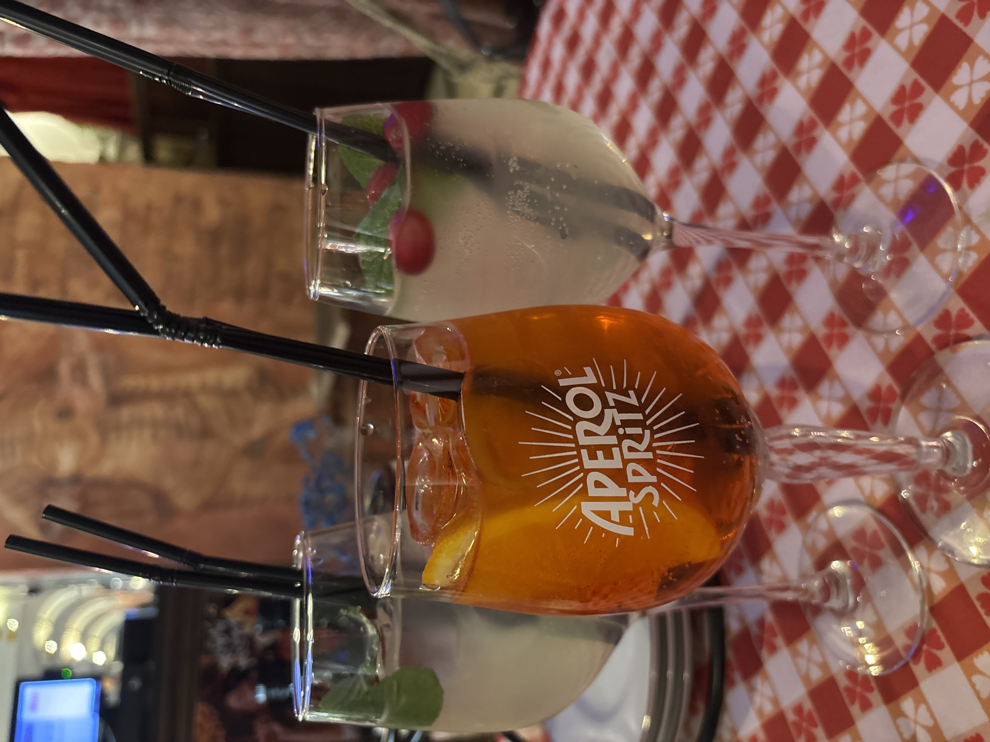 Three cocktails are displayed on a red and white checkered tablecloth featuring a subtle floral pattern, suggesting a restaurant or bar setting in Saint Petersburg, Russia. The ambient lighting indicates an indoor scene, likely in the evening.
The central drink is an Aperol Spritz, clearly identified by the prominent white "APEROL SPRITZ" logo, encircled by a sunburst design, on its glass. This bright orange beverage is garnished with an orange slice and contains a black straw.
Flanking the Aperol Spritz are two other cocktails served in similar stemware. The drink above is a lighter, possibly clear or cloudy beverage, garnished with fresh mint leaves and several red berries (possibly cranberries). The drink below is also clear or cloudy, garnished with mint leaves. All three glasses contain black straws. The background is blurred, showing hints of wooden textures and artificial lighting.
