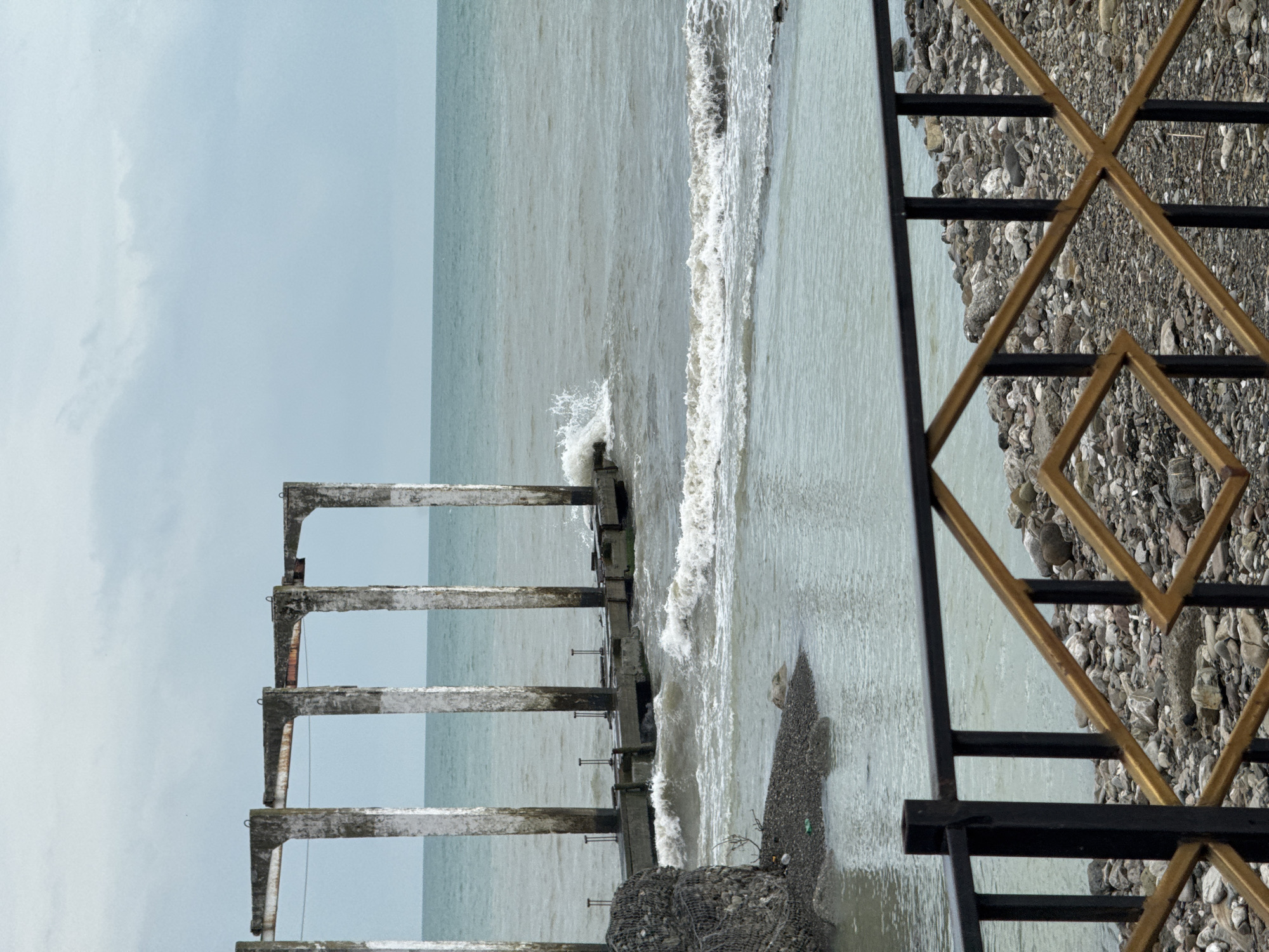 The photograph depicts a seaside scene near Sochi, Russia, taken from behind a decorative metal railing. The railing features a geometric pattern of gold-colored diamonds intersecting black vertical and horizontal bars. The foreground consists of a pebble beach, partially visible through the railing.
Beyond the railing, the scene opens to a view of the sea with gentle waves. A concrete structure, possibly the remnants of a pier or an old building, extends into the water. This structure consists of a series of eroded concrete pillars and beams, some of which are submerged in the water, and is damaged, showing visible signs of wear and age.
The sky is overcast, creating a soft, diffused light over the entire scene. The overall color palette is subdued, with grays, blues, and muted browns dominating the image. No people are visible in the frame.