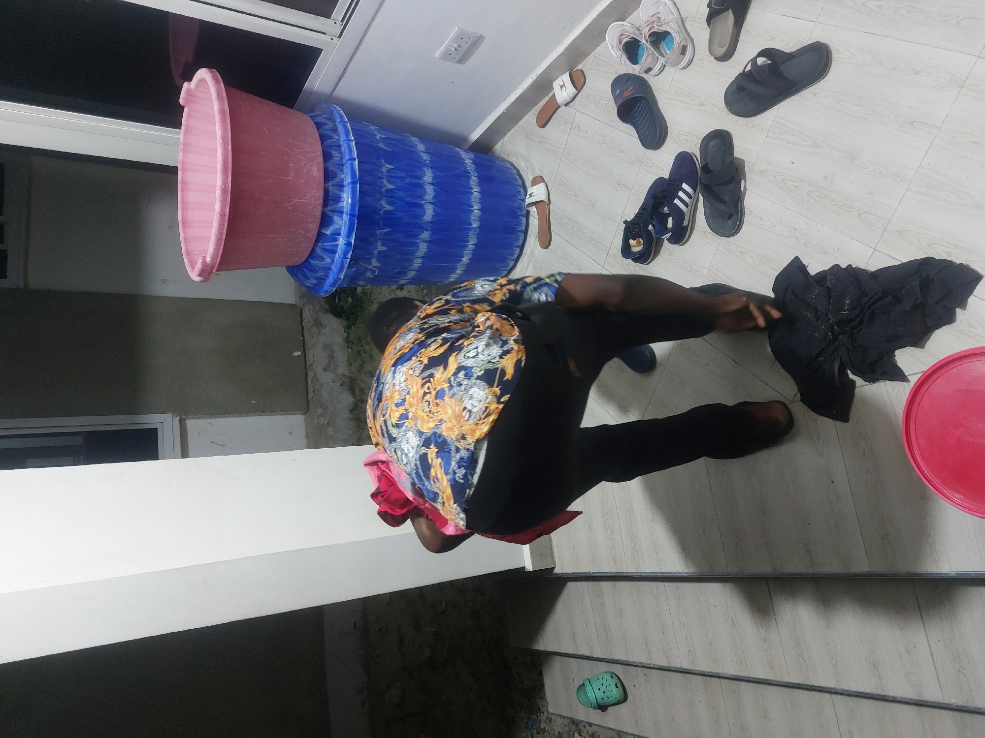 An overhead shot shows a man wringing out a dark cloth on a tiled floor near a collection of shoes. The floor also has a red lid near the man. In the background, stacked on top of one another are three plastic tubs colored pink and blue. The setting appears to be a porch or semi-outdoor area with a tiled floor. The location could be in Dar es-Salaam, Tanzania, based on general observations of the image, although it cannot be definitively confirmed.