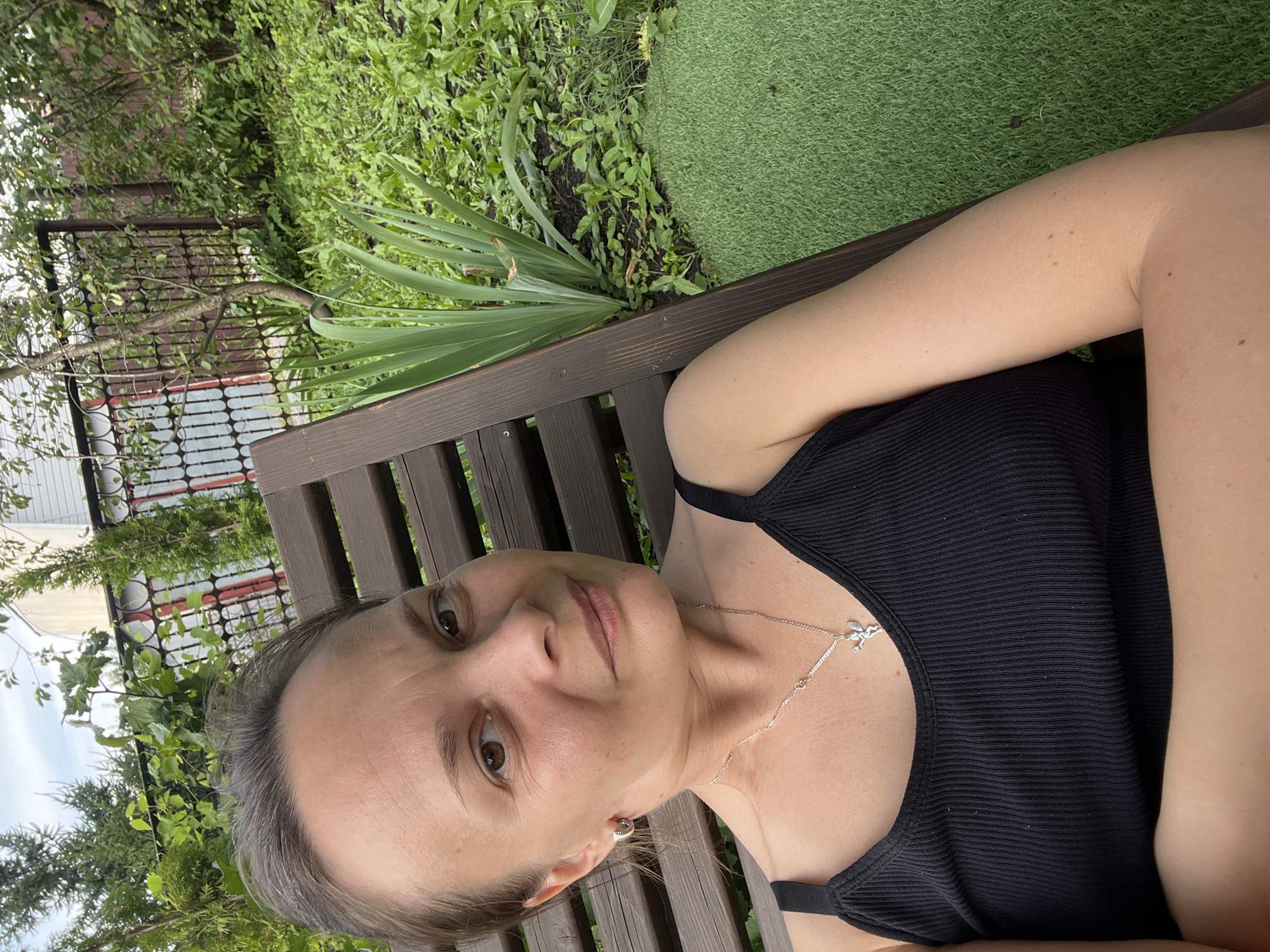 A woman is seated on a dark wooden lounge chair in a grassy yard. She is wearing a black tank top and a silver necklace with a small winged charm. Her arms are crossed. The background consists of lush green foliage, a dark metal fence, and a portion of a white building. The setting appears to be a residential backyard in Khryashchevka, Russia.