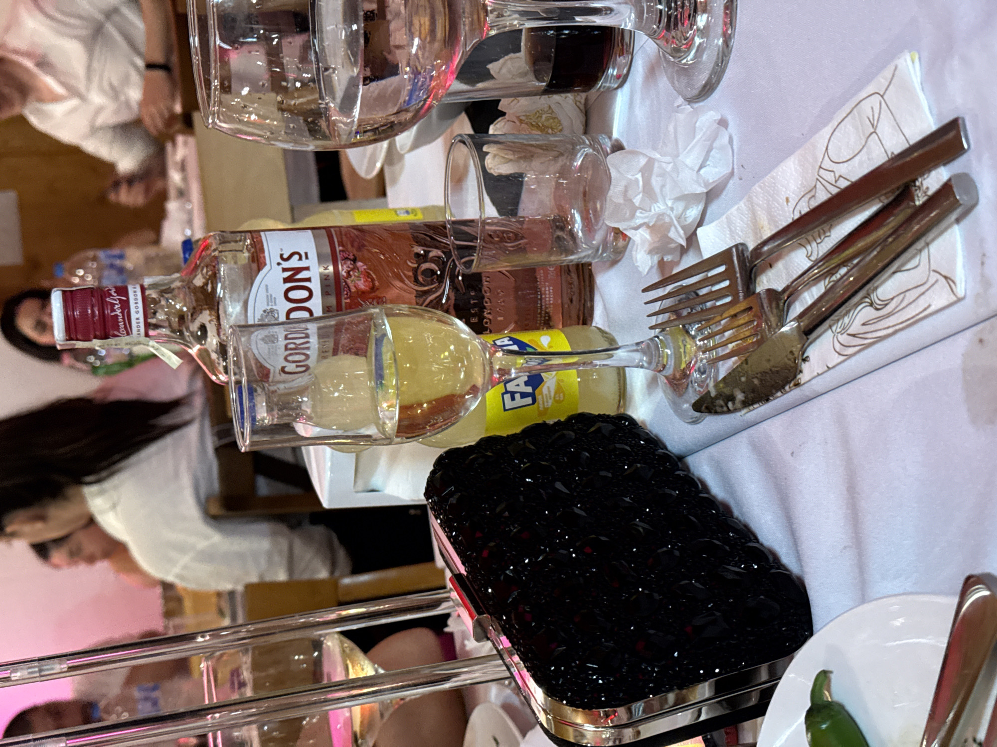 The image depicts an indoor dining or social gathering in Debrashtitsa, Bulgaria. The foreground features a white tablecloth with various items arranged upon it. A black, jewel-embellished clutch bag with silver-toned hardware is centrally positioned on the left. To its right, three pieces of silverware (two forks and one knife) rest on a white paper napkin adorned with a faint graphic of figures.
Multiple glasses and bottles are present on the table. A stemmed glass containing a yellowish liquid is visible on the left, partially obscured by two clear, vertical tubular elements, likely part of a table centerpiece. Next to this, a partial yellow bottle labeled "FANTA" is visible. A prominent bottle of "GORDON'S PREMIUM PINK DISTILLED GIN" with a red cap and pink liquid is centrally located. To the right of the gin bottle, an empty, clear, short tumbler glass sits beside a crumpled white napkin. Further right, a stemmed glass holds a dark liquid, and another stemmed glass contains clear liquid (water). A small section of a white plate with a green chili pepper is visible in the bottom-left corner.
The background is out of focus, revealing several individuals seated at tables. In the mid-left background, a person with dark hair, wearing a white collared shirt, is seated with their back partially to the camera. In the mid-right background, a male individual wearing a light-colored collared shirt is visible, with his hands positioned on the table. Other blurred figures and table settings are discernible throughout the background. The lighting in the background exhibits a pinkish hue, possibly from ambient illumination. A wooden chair back is visible in the mid-ground.