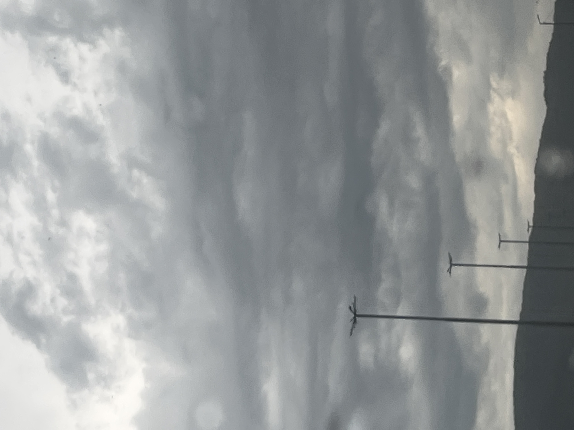 The media file depicts a scene with a cloudy sky and several tall poles with lights on them, spaced out in a line. The poles are silhouetted against the overcast sky. The sky itself is filled with a mix of dark and light gray clouds, suggesting a possibly stormy or overcast day. The overall mood is somewhat somber due to the weather conditions. The photograph is taken at an angle, making the sky the dominant feature. There are no visible people or distinct location cues in the image. The time of day is not clearly discernible, but the diffused light indicates it's not direct sunlight. There is no visible text in the image.