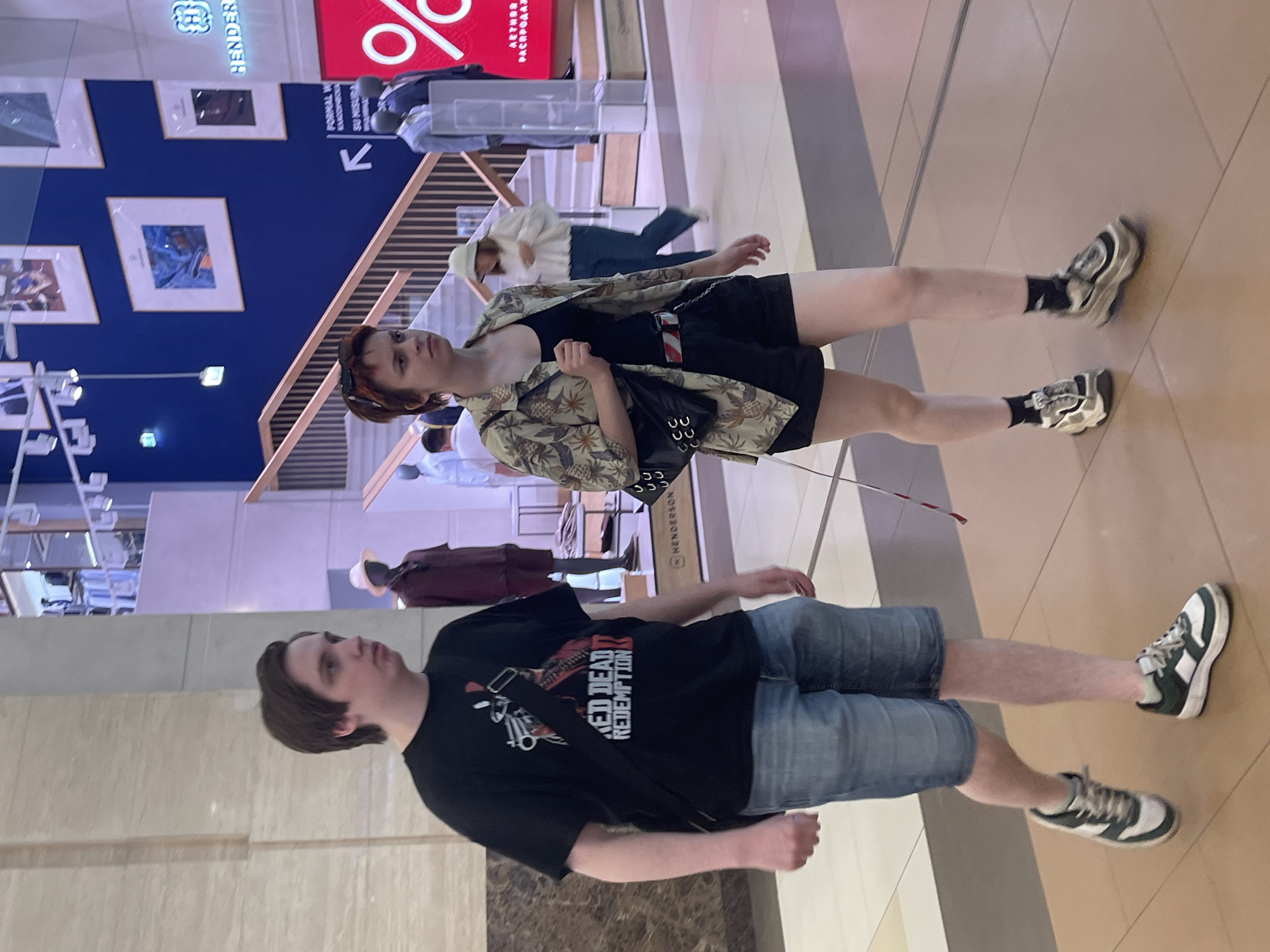 The image shows a young man with brown hair, wearing a black t-shirt with a "Red Dead Redemption 2" graphic, denim shorts, and sneakers, walking through an indoor shopping center. He has a black crossbody bag. Beside him, a young woman with short, reddish-brown hair and sunglasses on her head is wearing a patterned short-sleeved shirt over a black tank top, black shorts, and chunky sneakers. She is also carrying a black shoulder bag. In the background, a staircase with wooden railings is visible, and further back, mannequins dressed in formal wear and displays of clothing can be seen. There is also a large red sign with a "%" symbol and text in a foreign language. This appears to be a modern retail environment. The location is identified as Moscow, Russia, likely within a department store or mall.