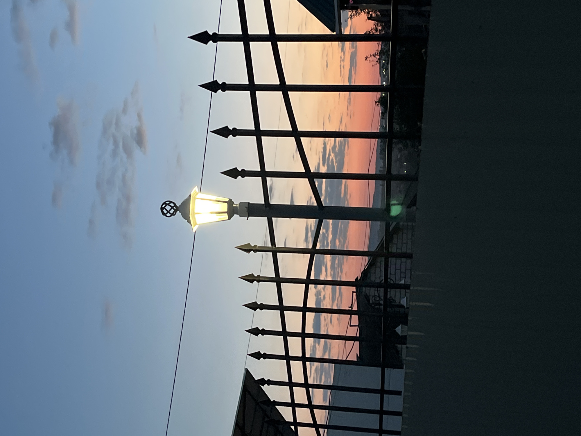 This image captures a dusky sky over a city, likely Saratov, Russia, with a silhouetted wrought-iron fence in the foreground. A vintage-style street lamp with a lit bulb is positioned centrally behind the fence, casting a warm glow. The sky transitions from a pale blue at the top to hues of pink and orange near the horizon, indicating either sunrise or sunset. Wispy clouds are scattered across the sky. Behind the fence, rooftops of buildings and some vegetation are visible, suggesting a residential or suburban area. The lower portion of the image is dominated by a dark, textured surface, possibly a wall or a gate. No people are present in the scene.
