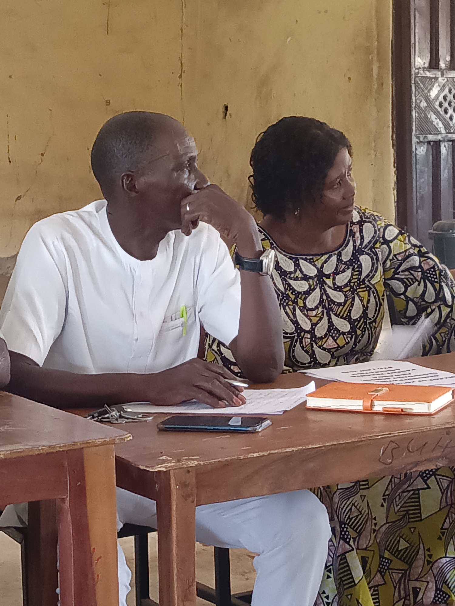 In an indoor setting, likely a classroom or meeting room in Akwanga, Nigeria, a man and a woman are seated at old wooden tables, appearing engaged in a discussion or listening intently.
The man, positioned on the left, has dark skin and closely cropped hair. He wears a short-sleeved white shirt with a mandarin collar, and a green pen is clipped into its left pocket. His left hand is raised to his chin in a thoughtful or pensive pose, and a ring is visible on his right hand. He is looking towards the right side of the frame.
To his right sits a woman with dark skin and dark, curly hair. She is wearing a vibrant, colorful patterned dress, possibly made of Ankara fabric. A watch is visible on her left wrist. Like the man, she is looking towards the right, appearing attentive.
The setting features distressed, light brown or yellowish walls, suggesting age. A window or door frame with a distinct geometric pattern is visible in the background. The wooden tables are old, showing signs of wear and tear. On the table in front of the man are some papers, a smartphone, and a set of keys. In front of the woman, there are more papers and a prominent orange notebook or planner with a clasp.
The overall atmosphere suggests an educational or community gathering. The visible text "B.M.H" is scratched into the side of the wooden table near the woman, and some illegible writing can be seen on the papers on the table. The lighting suggests it is daytime.