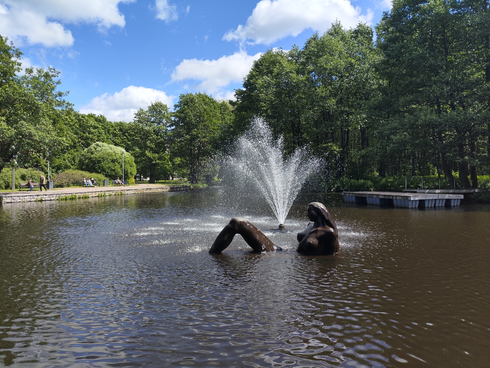 The image captures a serene view of a fountain in a body of water, likely a pond or lake, within a park setting. The fountain is centrally located, with water arcing upwards from its center, creating a misty spray. A dark-colored sculpture of a figure, partially submerged in the water, is visible in the foreground, with one leg extending out of the water at an angle and the torso and head of the figure facing to the right. The water is a deep brown color, reflecting the surrounding trees and sky. Lush green trees form a dense backdrop, with their branches reaching towards the bright blue sky dotted with fluffy white clouds. In the background, on the left side of the image, a paved walkway lines the edge of the water, with several people seated on benches and others walking. To the right, a wooden deck structure is situated on the water's surface. This scene is located in Kadriorg Park in Tallinn, Estonia, specifically the pond area near the Kadriorg Palace.