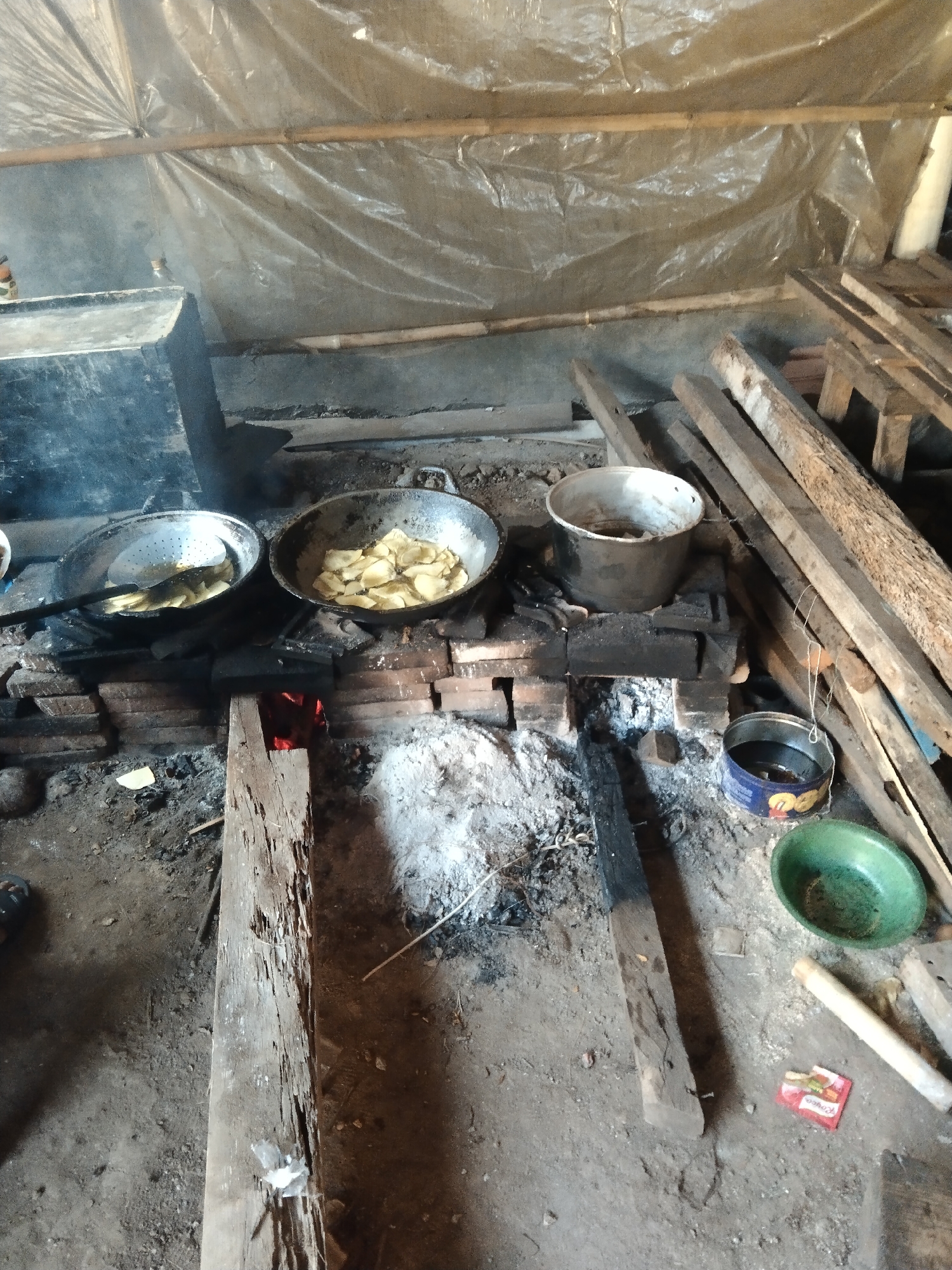 The image depicts an outdoor cooking area in Kelet, Indonesia. A primitive stove constructed from bricks is visible, with active embers glowing within its firebox. Two pans are placed on top of the stove. One pan, on the left, appears to contain oil and food being drained by a slotted utensil. The other pan, on the right, is filled with what looks like sliced potatoes or plantains being fried. Adjacent to the stove is a large metal container, possibly used for storage. To the right of the stove, a collection of rough wooden planks are stacked. On the ground, near the stove, are a small tin can, a green bowl, and a red packet of what appears to be spices or instant noodles. The ground is covered in dirt, ash, and debris. The background is obscured by a translucent plastic sheeting supported by bamboo poles, suggesting a temporary or rudimentary shelter. The overall scene conveys a sense of basic, functional outdoor food preparation.