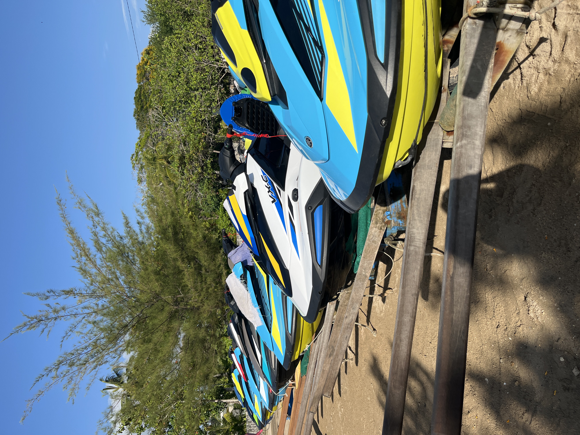 In Kuta Selatan, Indonesia, a line of colorful jet skis is parked on a sandy beach under a clear blue sky. The jet skis are predominantly white, blue, and yellow, with some featuring black accents. They are positioned on wooden stands and appear to be ready for rental. Behind the jet skis, lush green trees and vegetation form a backdrop, with a tall, slender tree in the foreground. The scene conveys a sense of a popular tourist destination, likely a beach resort.