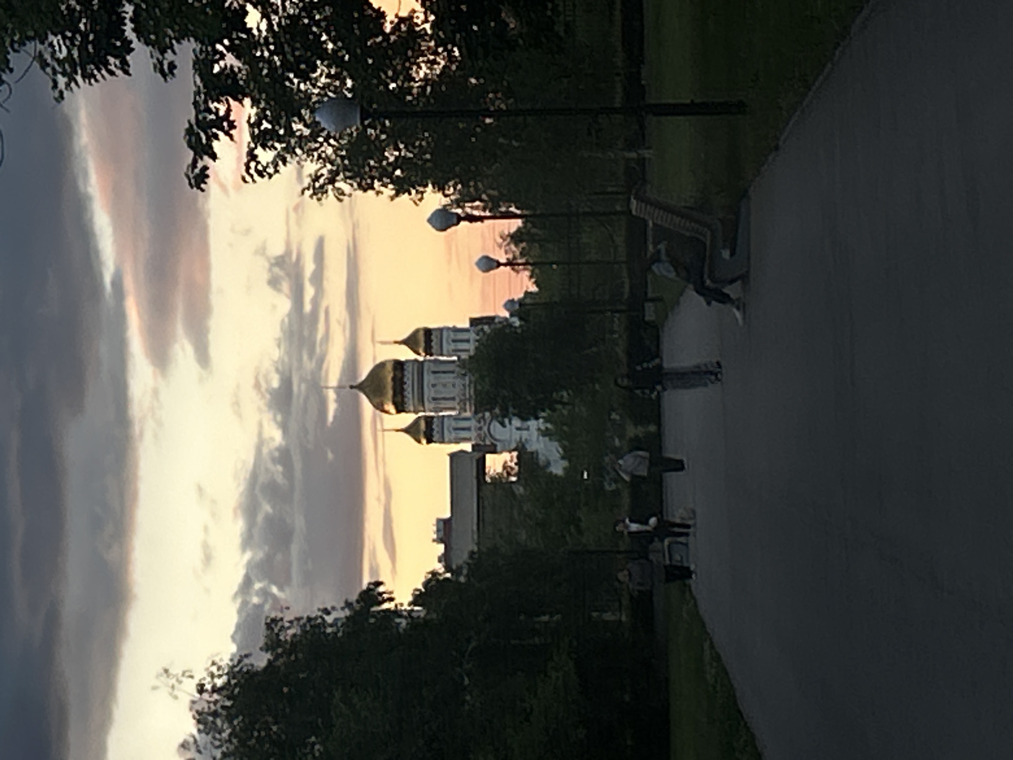 The image shows a view of a cityscape at dusk. Golden domes of a cathedral are visible in the distance, framed by trees. A row of streetlights lines a pathway on the right side of the frame, where several people are walking or sitting. The sky displays a mix of dark gray and pastel colors, suggesting a sunset. The location is possibly in a park in Tolyatti, Russia.